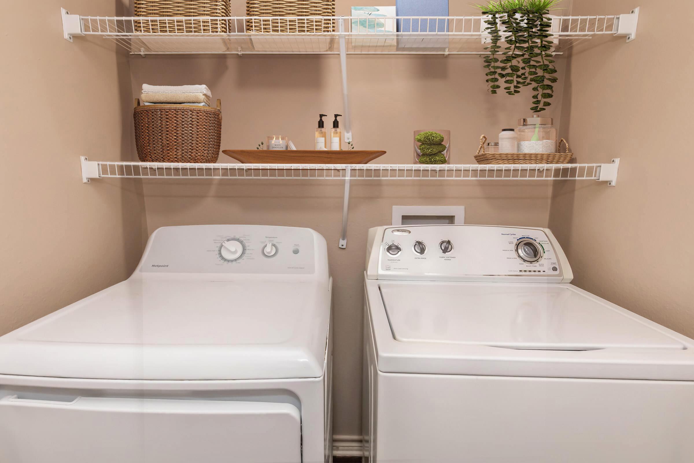 A laundry room featuring two white washing machines side by side. Above them, there are shelves with woven baskets, neatly arranged towels, and small plants for decor. The walls are painted a soft neutral color, creating a clean and organized space.