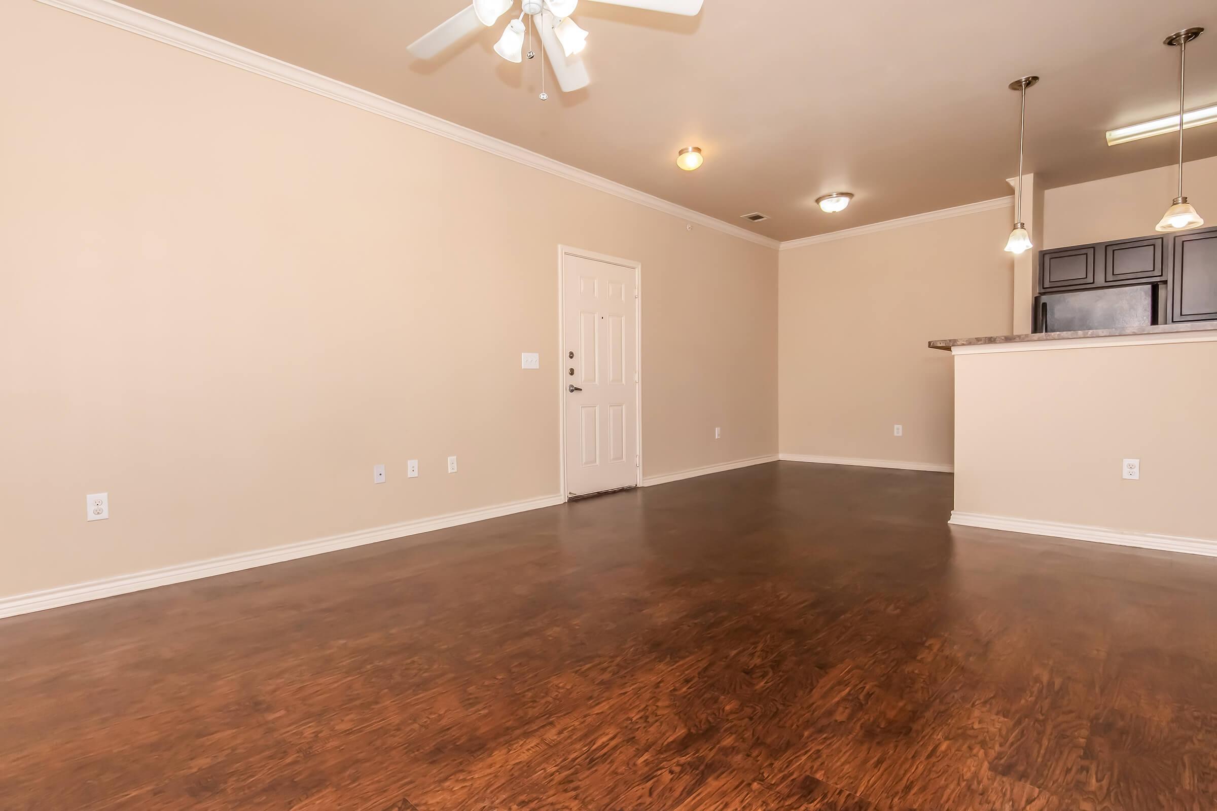 A spacious, empty living room with light beige walls and dark wood flooring. There's a ceiling fan with lights, a white door leading to the hallway, and an open view towards a kitchen with a bar counter. The overall ambiance is bright and inviting, creating a blank canvas for decor.