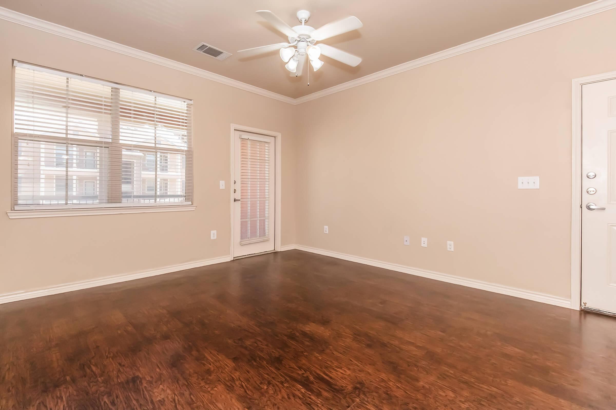 Spacious, empty living room featuring a light-colored wall, hardwood flooring, a ceiling fan with light, and a large window with blinds. A glass door leads to a balcony, and the entrance is visible on the right side. The room has a clean, modern aesthetic.