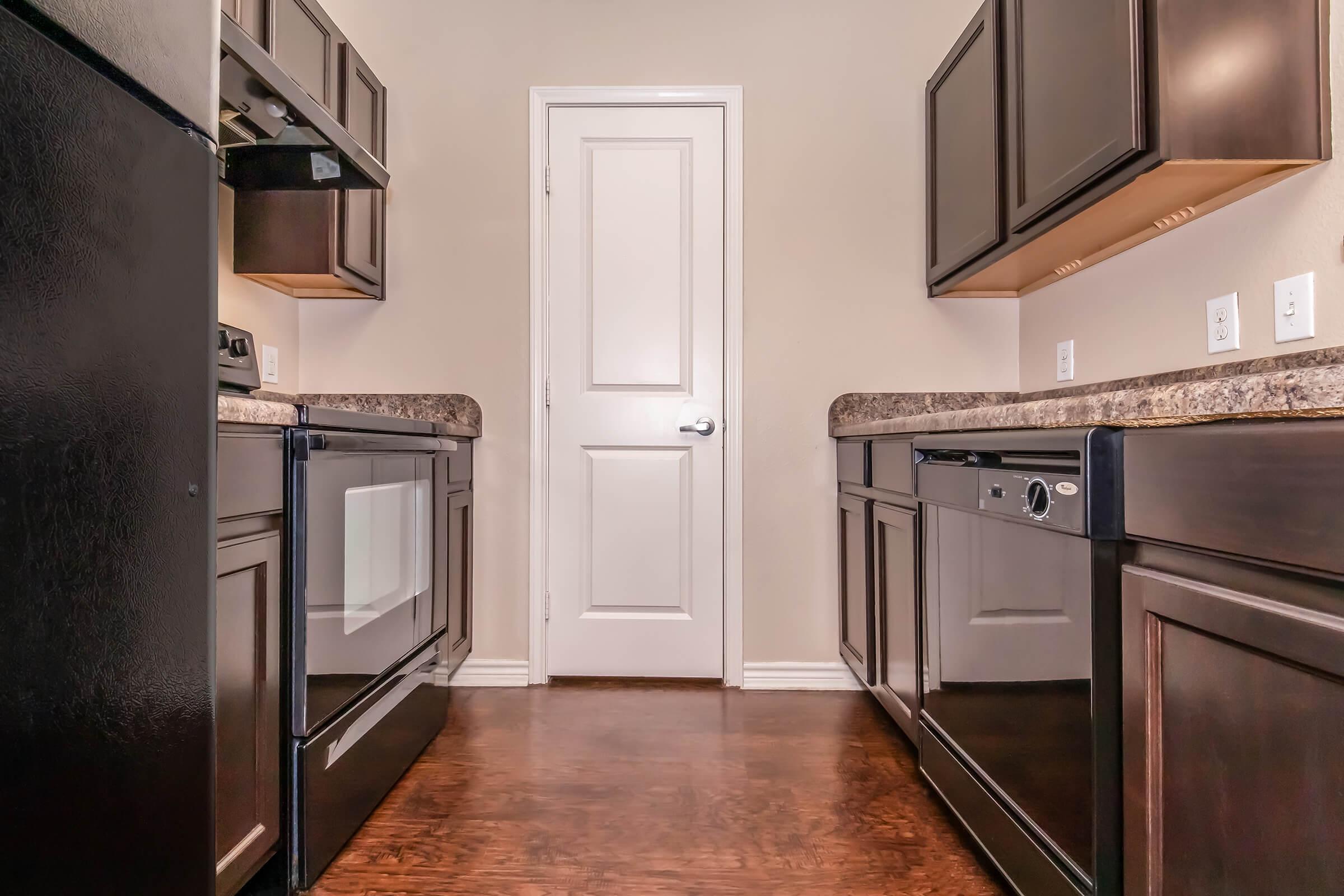 A modern kitchen interior featuring dark wooden cabinets, granite countertops, and black appliances including a refrigerator, oven, and dishwasher. The space has a door at the far end and wooden flooring, creating a warm and inviting atmosphere.
