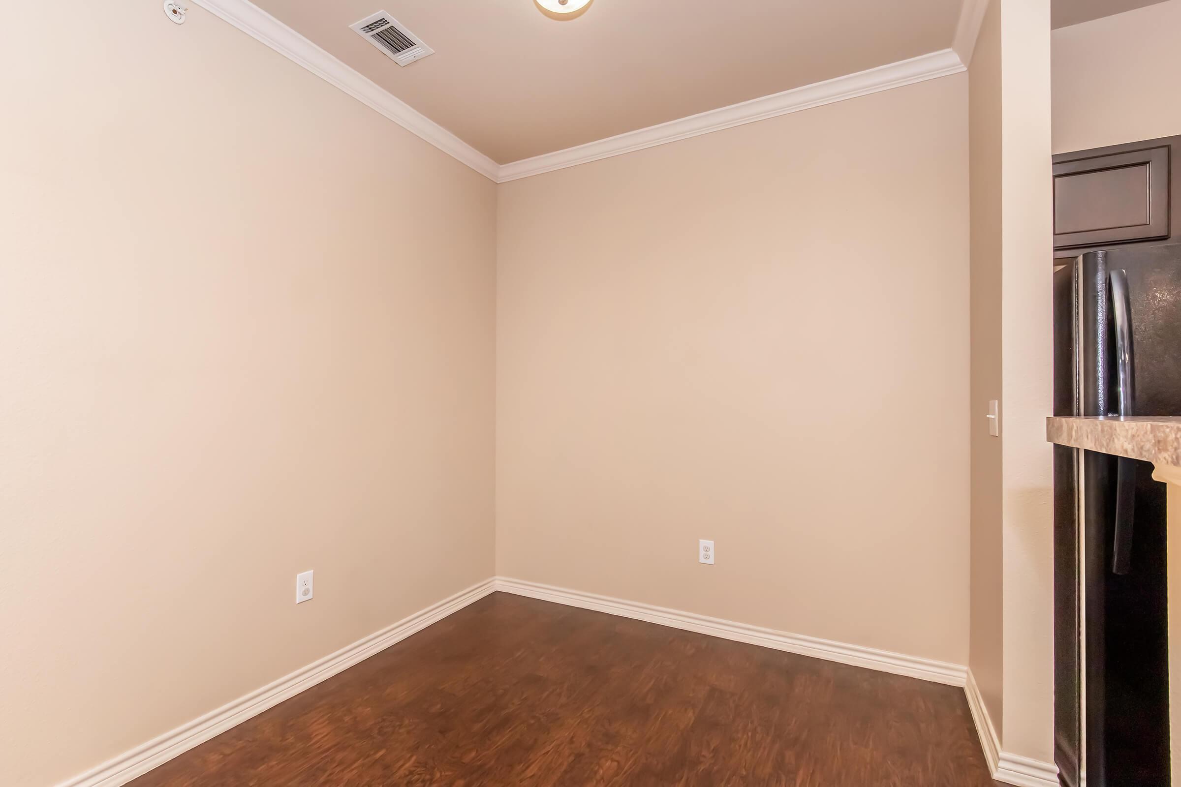 Empty room with light beige walls and wooden flooring. There are no furniture items present. A corner features a black refrigerator, while the ceiling has a light fixture. The overall space appears clean and well-maintained, suitable for various uses.