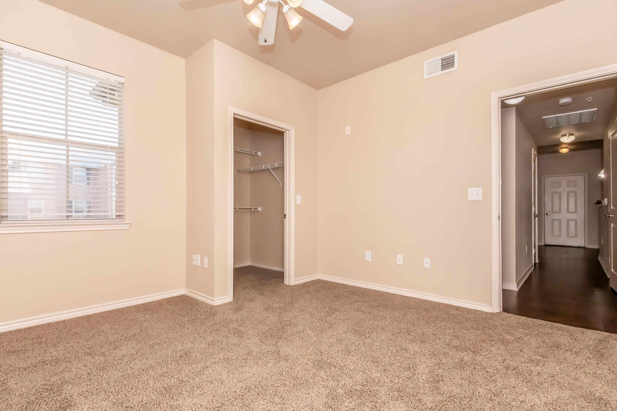 A spacious room with tan walls and carpet, featuring a ceiling fan. There is a window with blinds allowing natural light, and a closet on one side. The area has a smooth transition to a hallway visible in the background, which leads to other rooms. The overall atmosphere is warm and inviting.