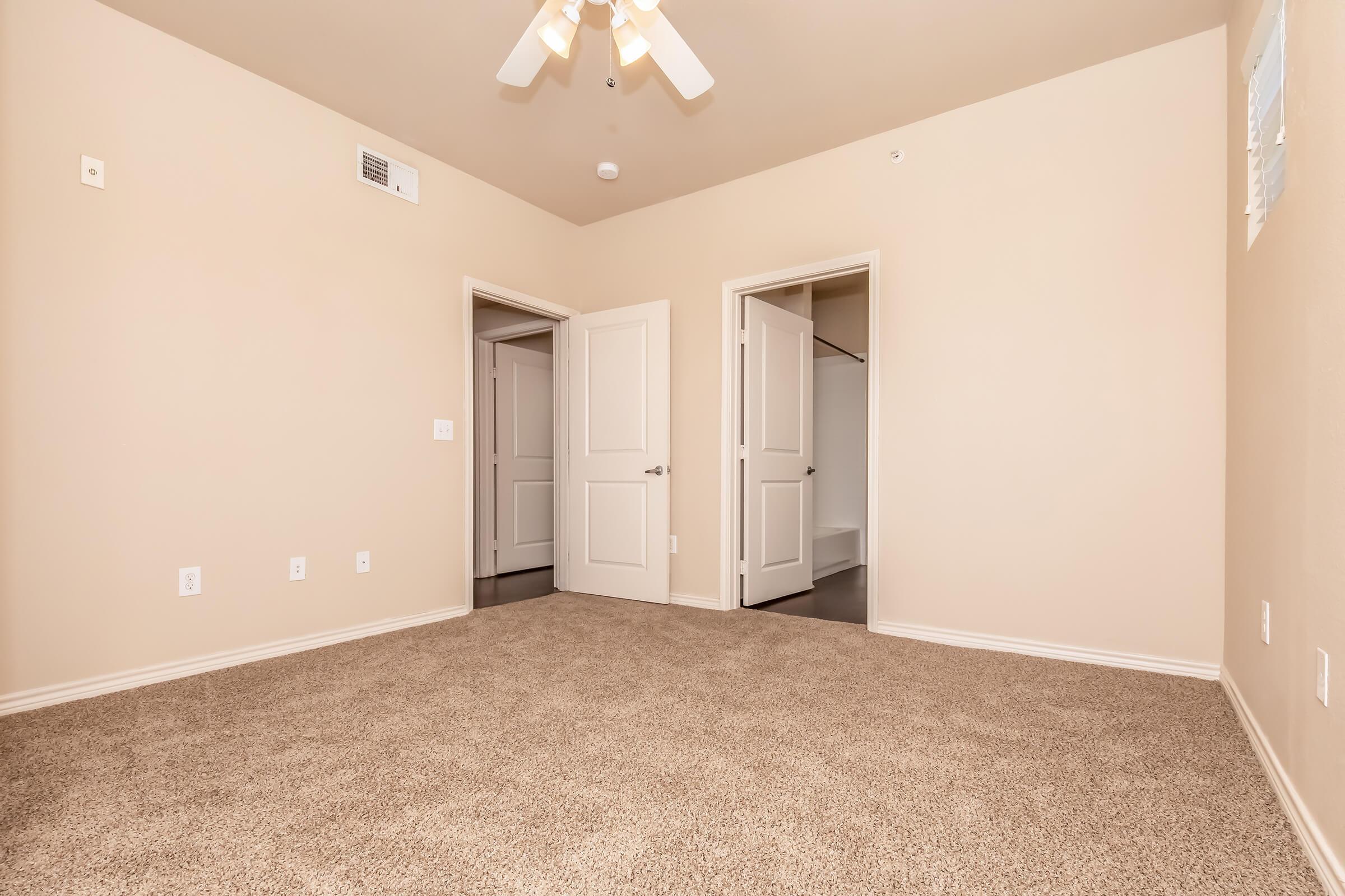 A spacious, neutral-toned bedroom with light beige walls and plush carpeting. The room features a ceiling fan, two doorways leading to closets, and no visible furnishings. Natural light enters through the doorways, creating a warm and inviting atmosphere.