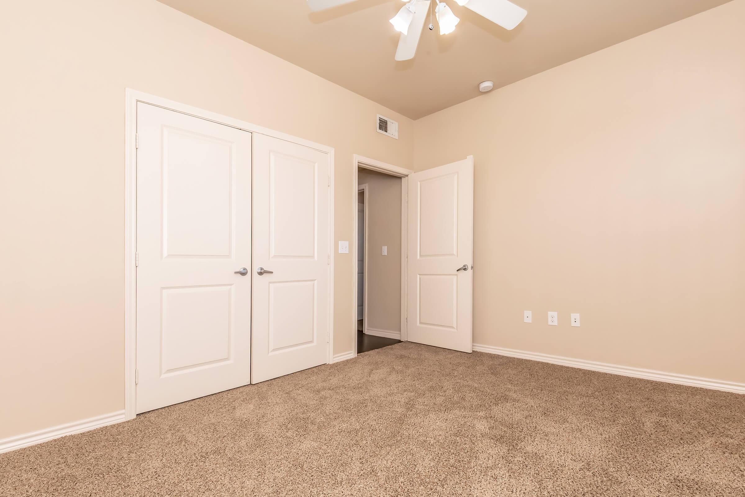 A spacious, empty room featuring beige walls and carpet, with a ceiling fan. There are two white double doors on one side and an open doorway leading to another room. The lighting is bright, showcasing the clean and uncluttered space, perfect for personal customization.