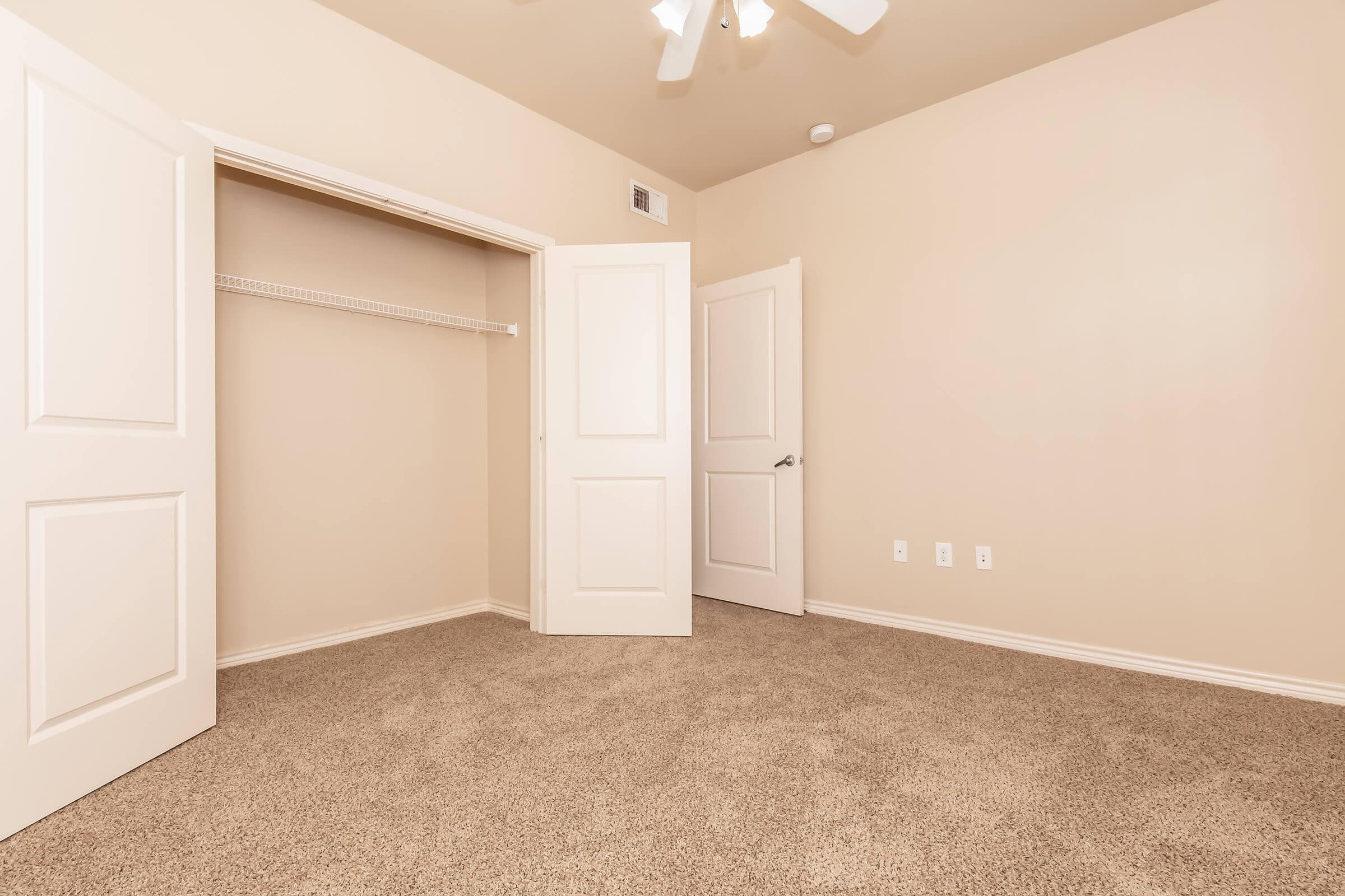 An empty bedroom featuring light beige walls and carpet. There are two open white closet doors on the left revealing a shelf inside, while another door leads to a different room on the right. A ceiling fan is visible above, and the overall space is bright and uncluttered.