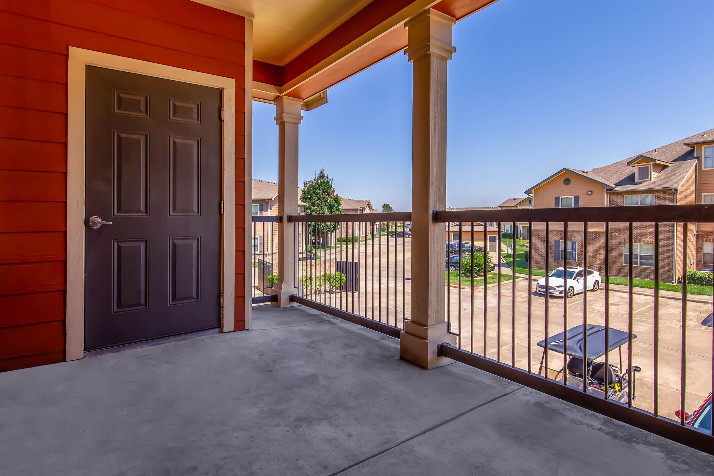 A spacious balcony view featuring a closed dark door, concrete flooring, and a railing. Beyond the railing, there are residential buildings, parking spaces with cars, and clear blue skies. The scene suggests a pleasant, sunny day in an apartment community.