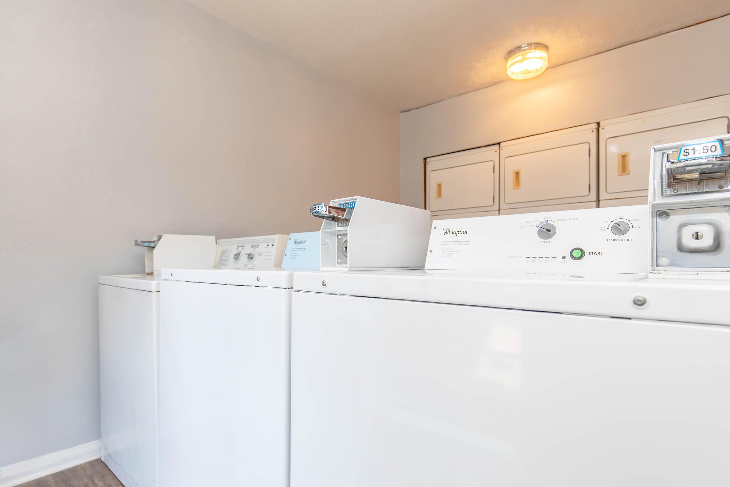 A clean laundry room featuring several white washing machines and dryers lined up against a light gray wall. A bright overhead light illuminates the space, highlighting the laundry machines and their controls. The floor is a light wood finish, adding to the bright, tidy atmosphere.