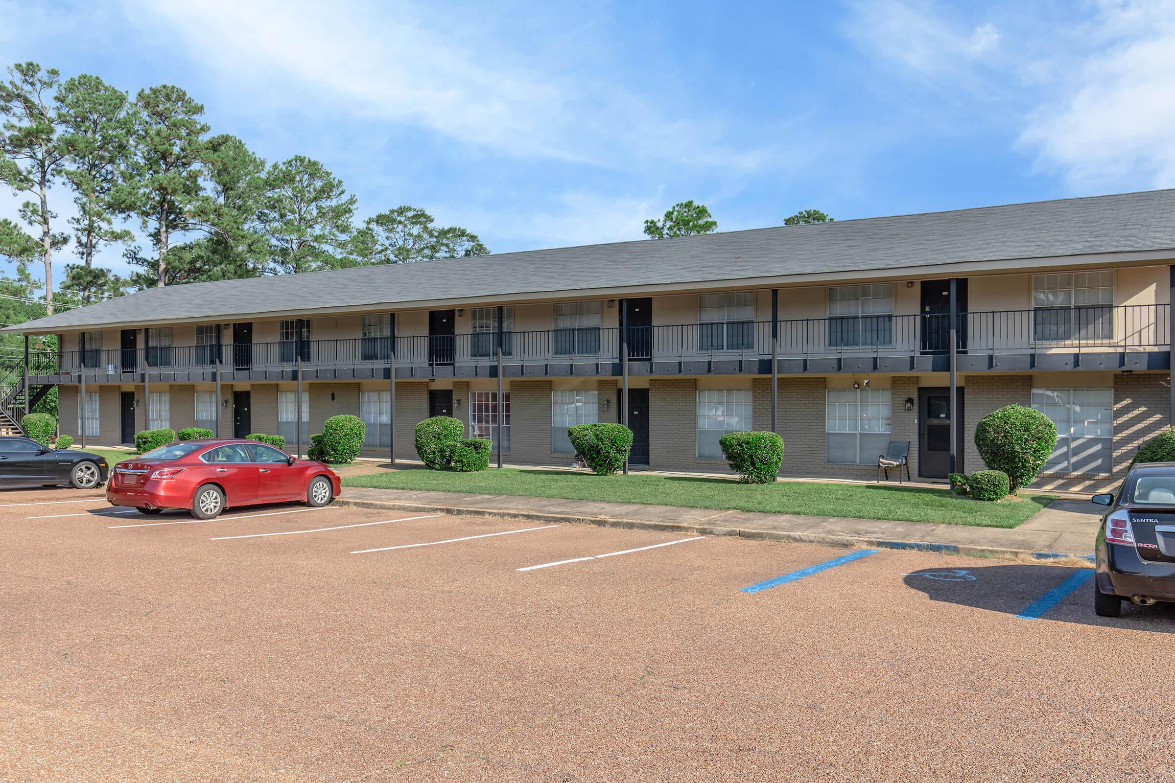 Two-story motel with a row of rooms, each featuring balconies. The exterior is light-colored brick with dark shutters. Neatly trimmed bushes line the front, and several parked cars, including a red one, are visible in the parking lot. The sky is blue with a few clouds.