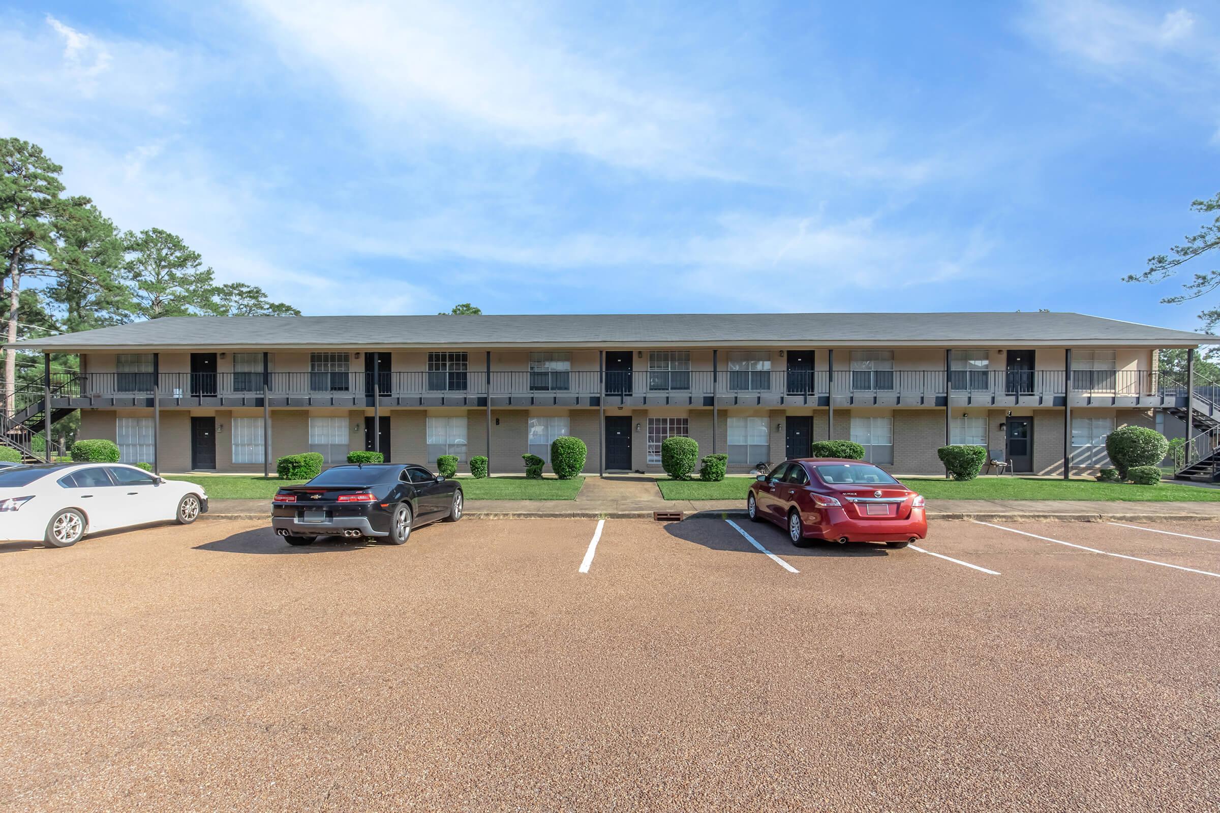 A two-story apartment complex with a beige exterior and black railings. There are several parked cars in the foreground, including a white sedan, a black sedan, and a red sedan. The area is surrounded by greenery, with trees visible in the background under a clear blue sky.