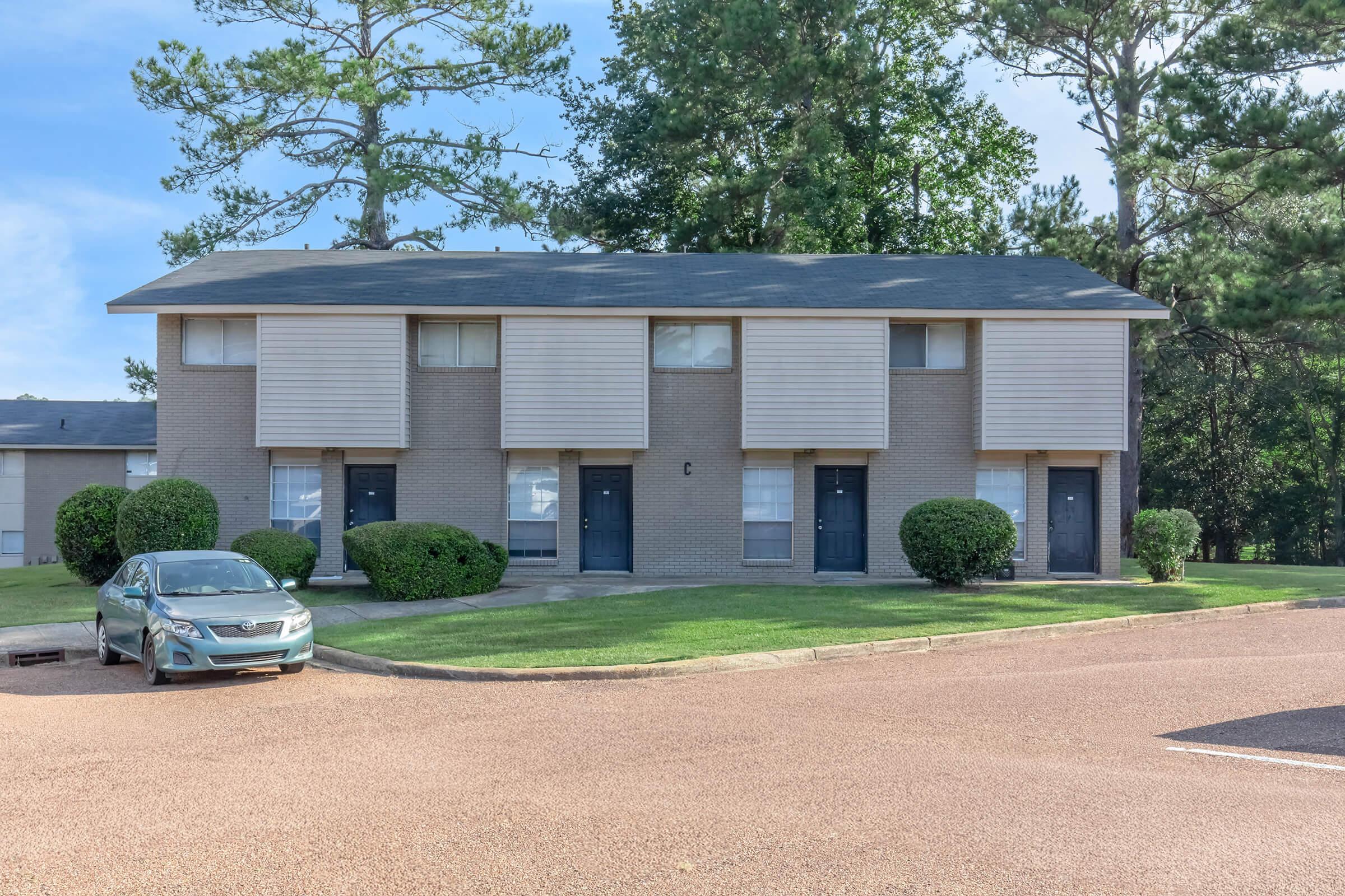 A two-story apartment building features multiple units with light brown brick walls and shuttered windows. The lawn is well-maintained, with shrubs planted in front. A parked silver car is visible near the entrance, and there are trees in the background under a blue sky.