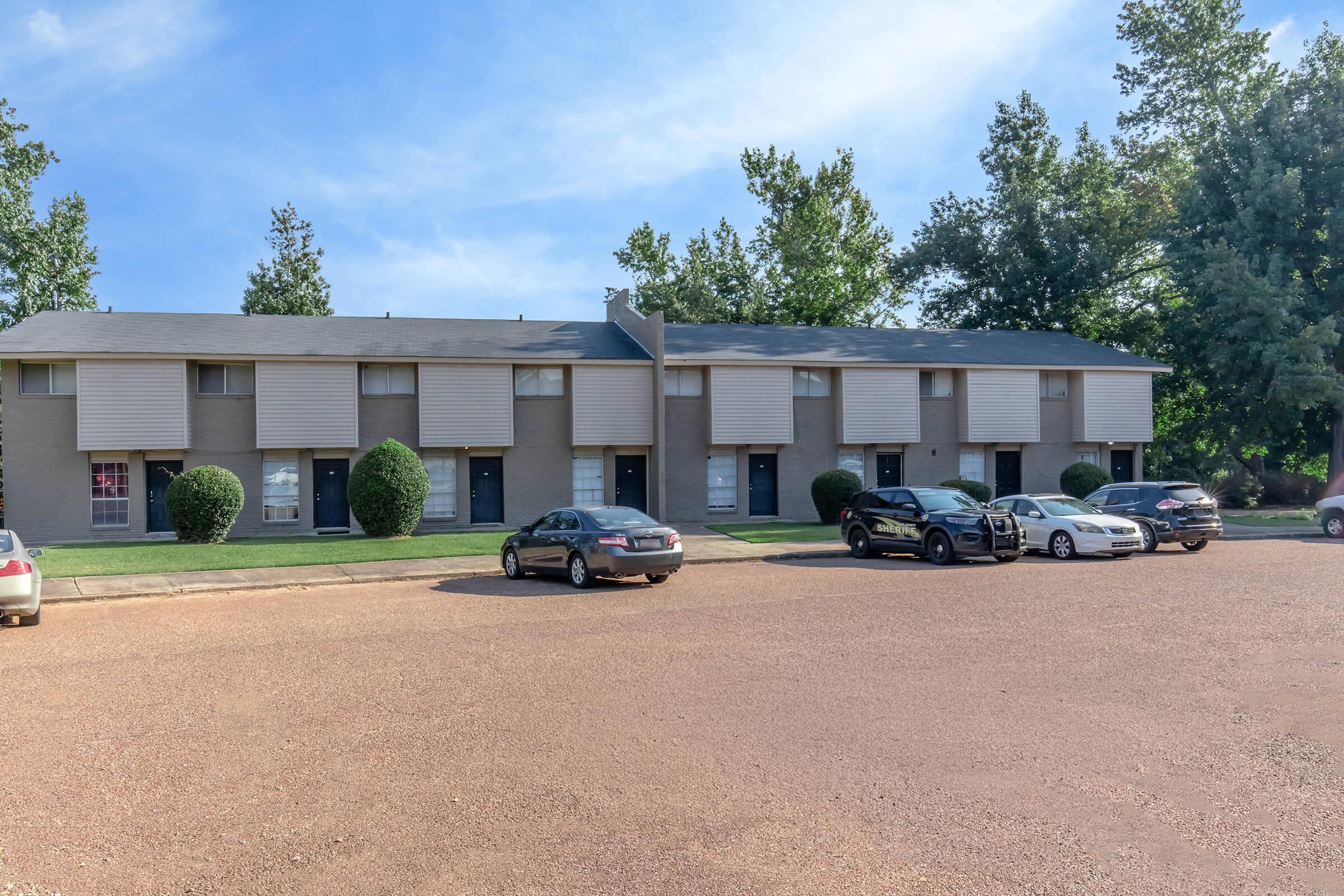 A row of two-story apartment buildings with beige siding and shuttered windows. The parking area in front has several parked cars, surrounded by well-maintained grassy areas and trees in the background, under a clear blue sky.