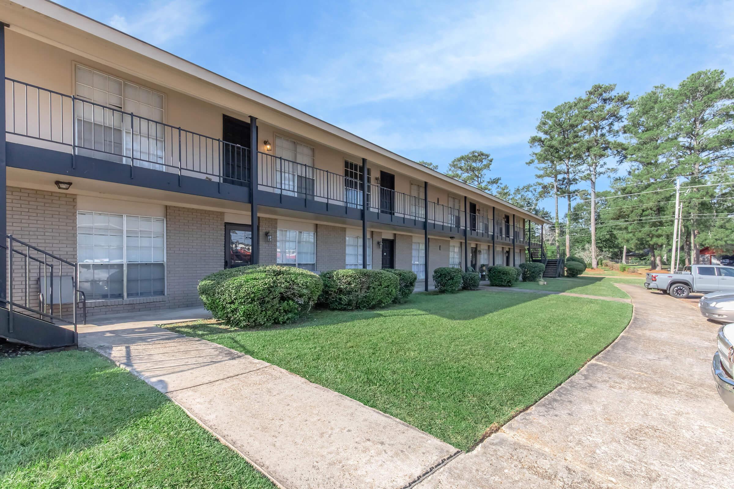 Two-story apartment building with a row of balconies on the second floor. The building features multiple windows and is surrounded by well-maintained green lawns and shrubs. A concrete pathway leads to the entrance, and there are parked cars nearby under a clear sky.
