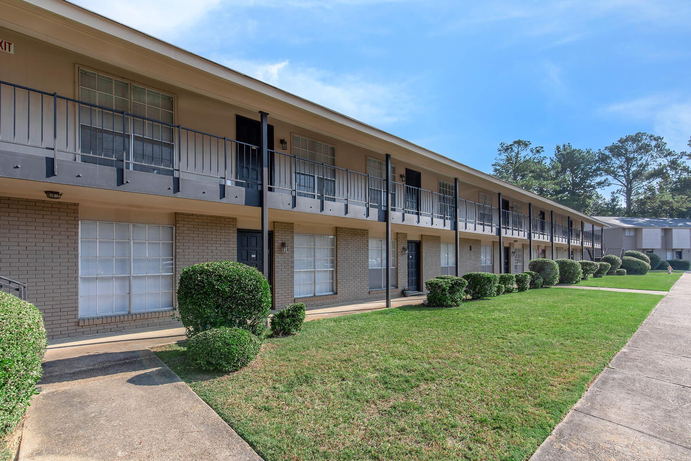 A row of multi-story apartment units with balconies, surrounded by neatly trimmed bushes and a well-maintained lawn. The sky is clear and blue, and the building features a neutral color scheme with large windows. A concrete pathway leads up to the entrance of the apartments.