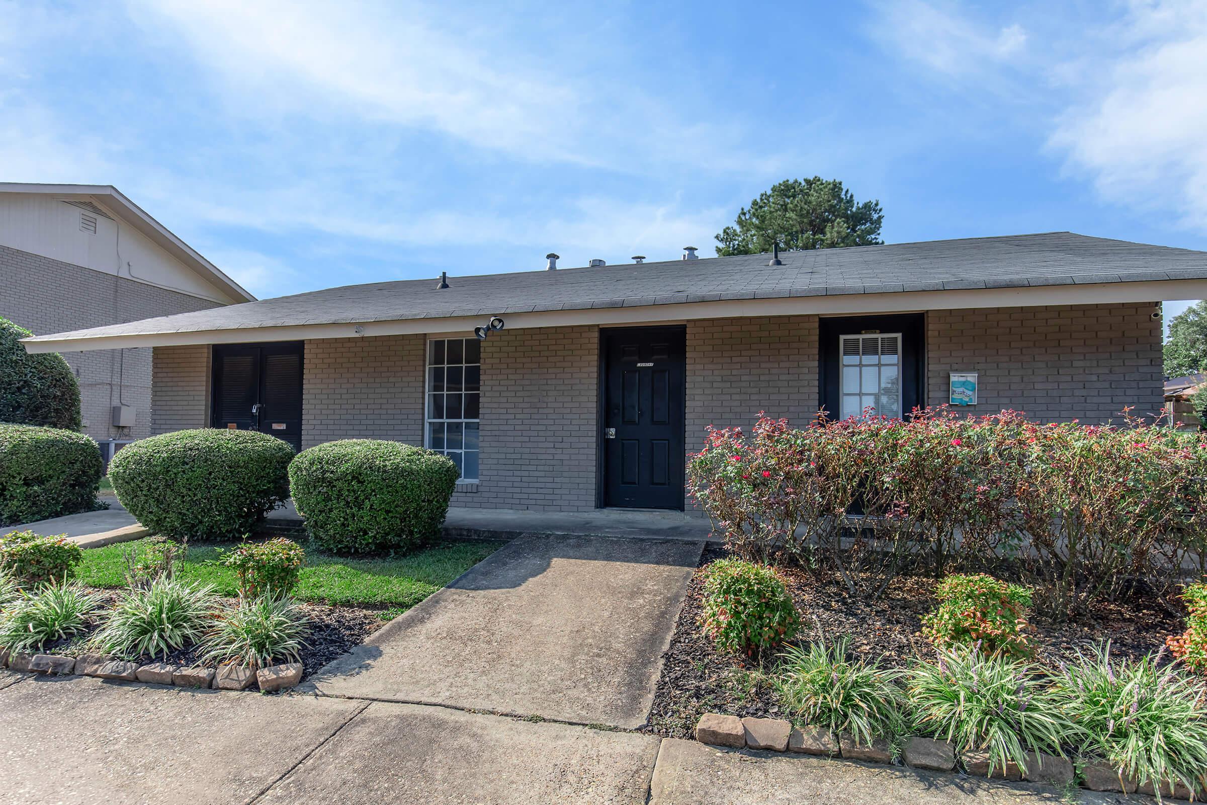 A single-story brick house featuring a black front door, two windows on either side, and neatly trimmed shrubs in the front yard. A concrete pathway leads to the entrance, surrounded by colorful landscaping. The sky is clear with a few clouds visible above.