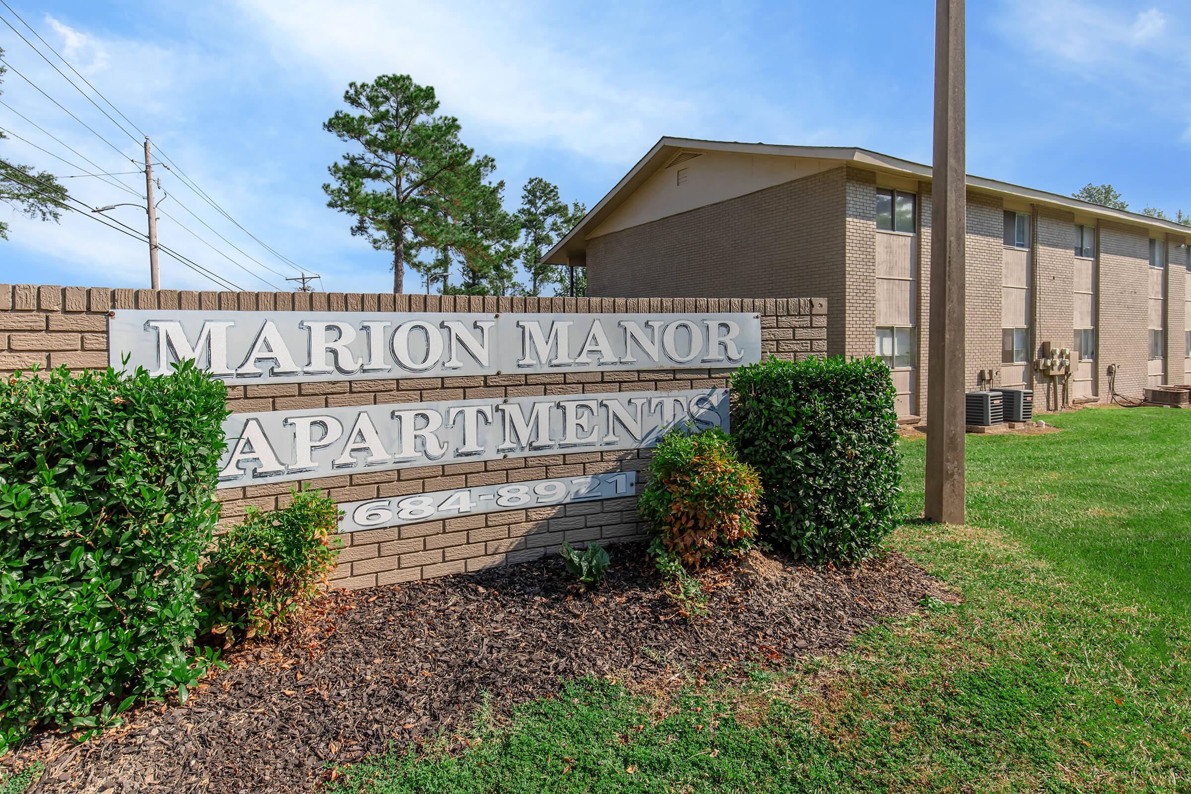 Sign for Marion Manor Apartments, featuring the name prominently displayed on a brick wall. The sign is surrounded by well-maintained landscaping, including bushes and grass. In the background, a two-story apartment building is visible, along with utility poles and trees.