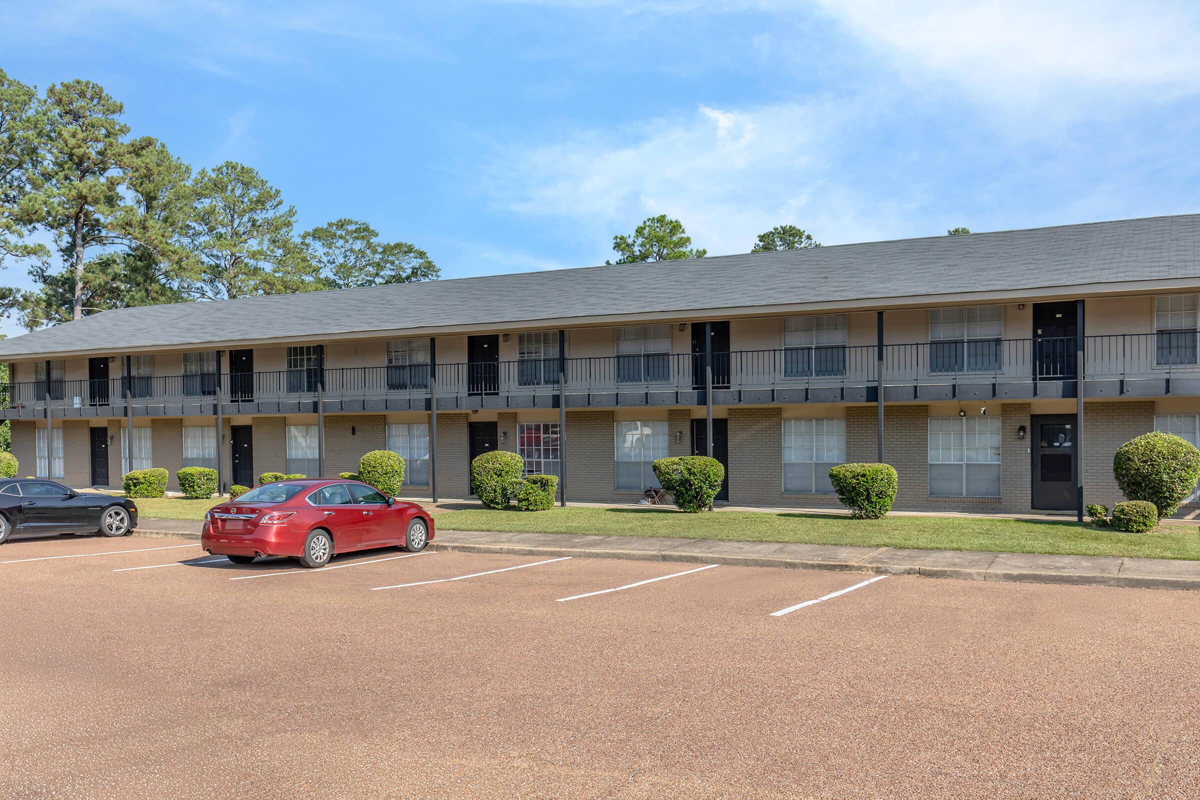 A two-story residential building with multiple units, featuring balconies and large windows. In front, there are two parked cars—a red one and a black one—on a gravel parking lot. The area is landscaped with small hedges and trees, under a clear blue sky.