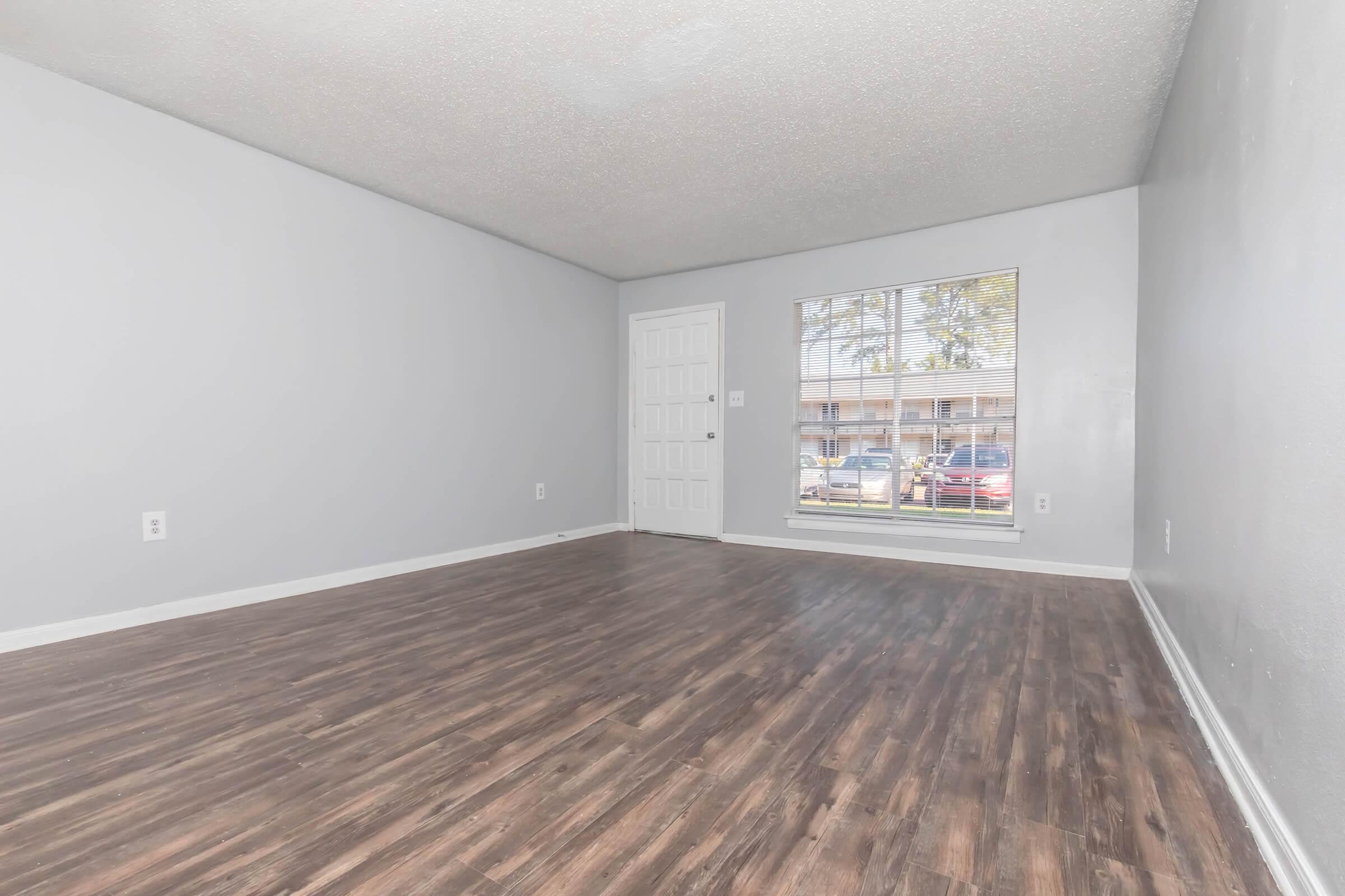 Empty room with light gray walls and a dark wooden floor. A large window with blinds lets in natural light, showcasing a view of the outside. There is a white door on the left side of the wall, and the space appears clean and ready for furnishing.