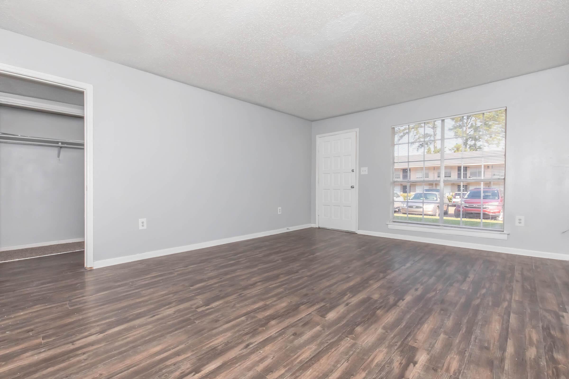 A spacious, empty room with light gray walls and dark wooden flooring. A window lets in natural light, revealing an exterior view. On the left, there's an open closet space. The overall atmosphere is clean and modern, suitable for various interior designs.