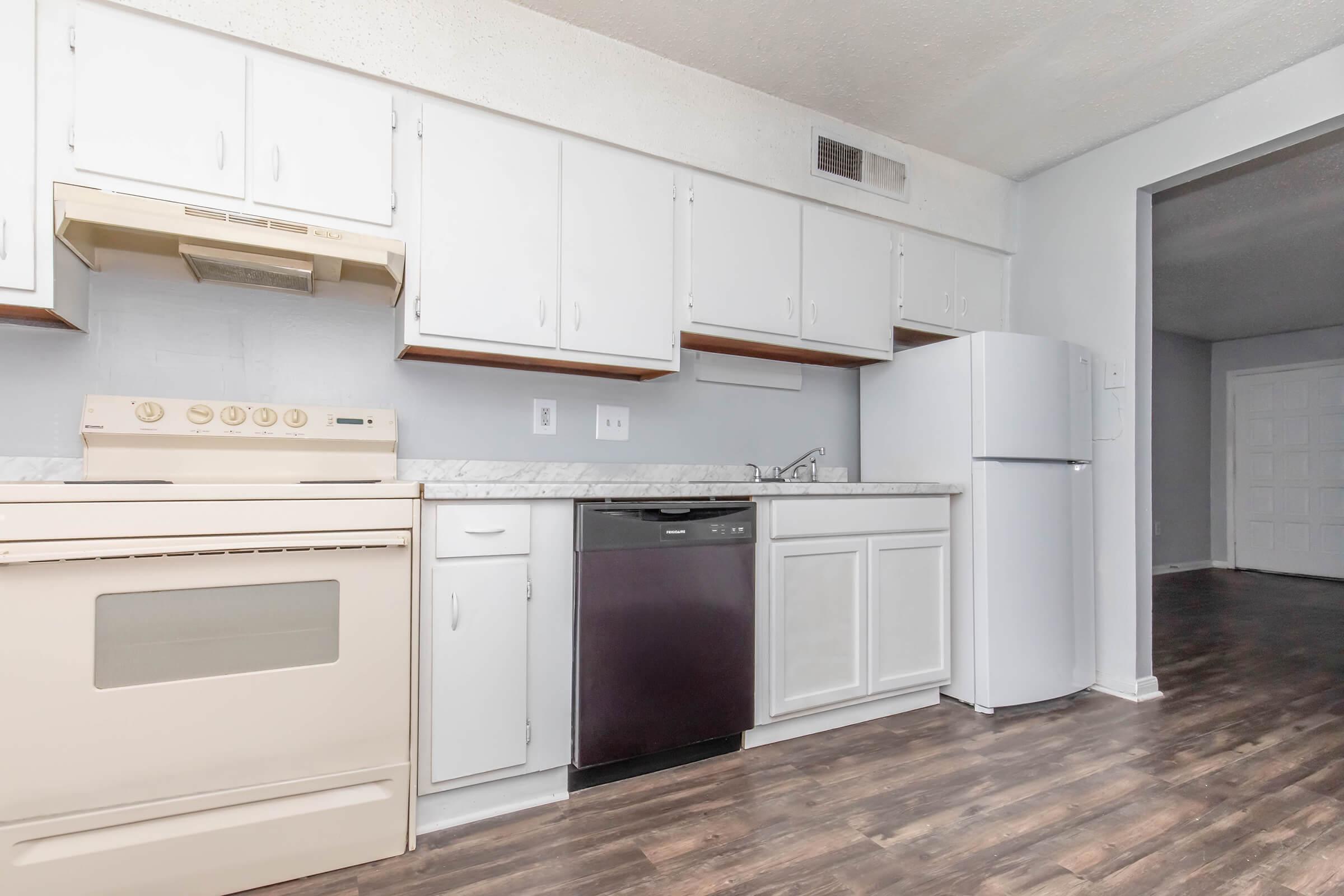 A modern kitchen featuring white cabinetry, a light-colored countertop, a built-in oven, and a dishwasher. A refrigerator stands nearby, with a view into a larger room beyond. The flooring is dark wood, creating a warm contrast with the bright kitchen elements.