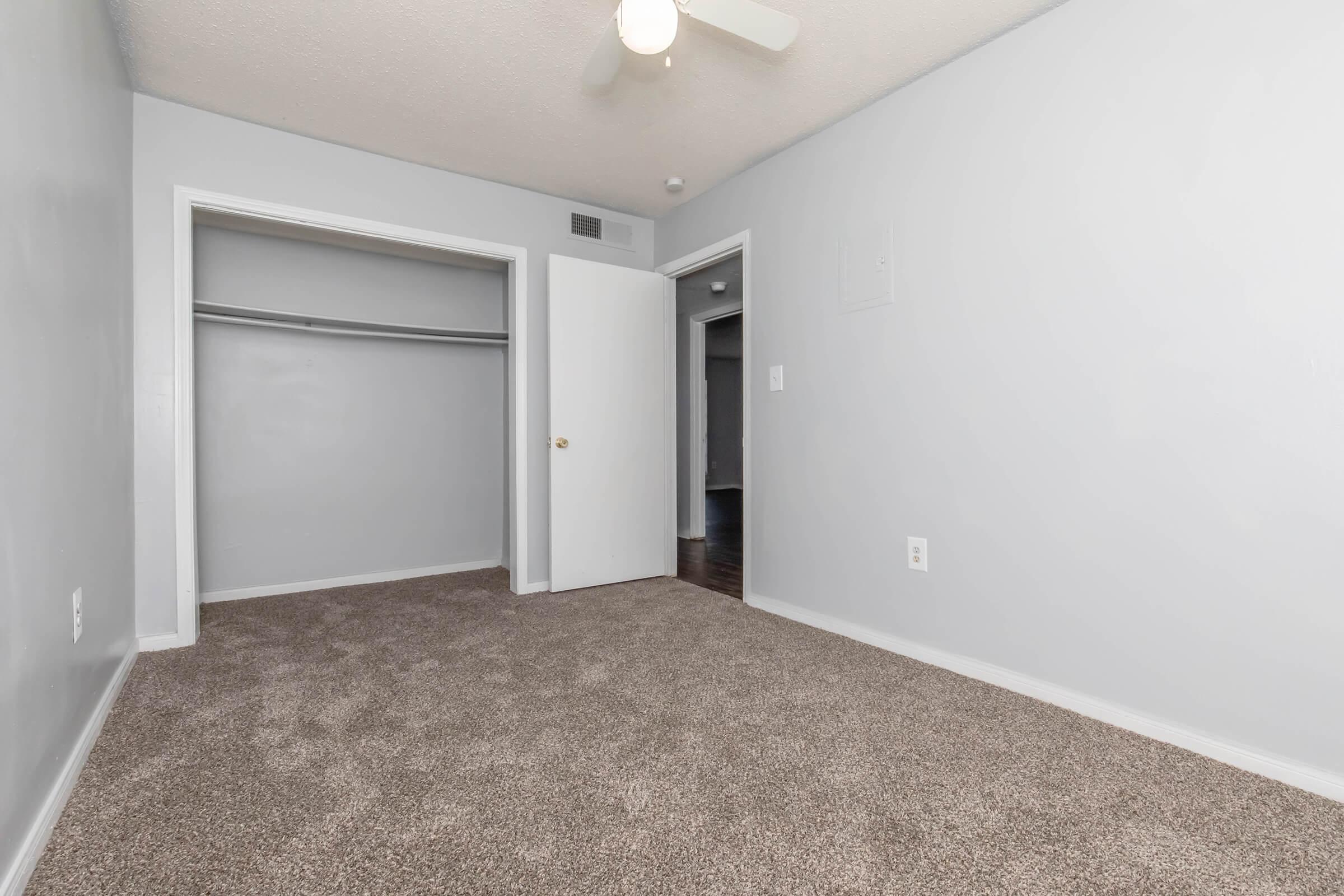 A clean, light gray bedroom featuring carpet flooring. The room includes a ceiling fan and two doors: one leading to a closet and another to a hallway. The walls are painted in a soft shade, creating a spacious and inviting atmosphere.