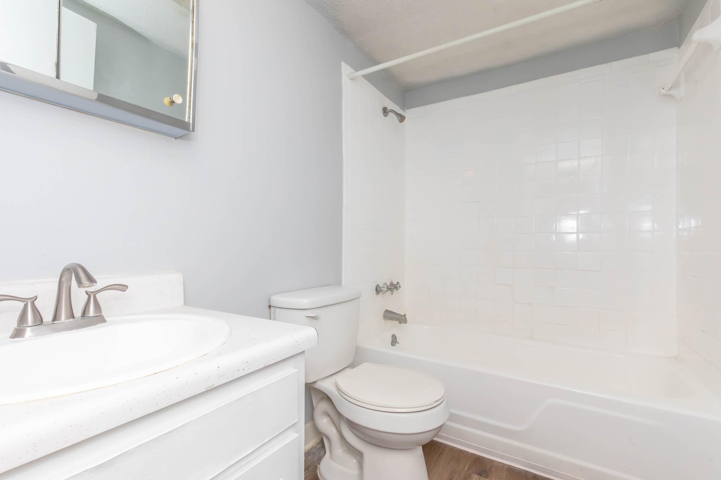 A clean, modern bathroom featuring a white bathtub and shower combination, a white toilet, and a vanity with a sink. The walls are painted light gray, and there is a mirror above the sink. The flooring appears to be a wooden design, enhancing the overall aesthetic of the space.