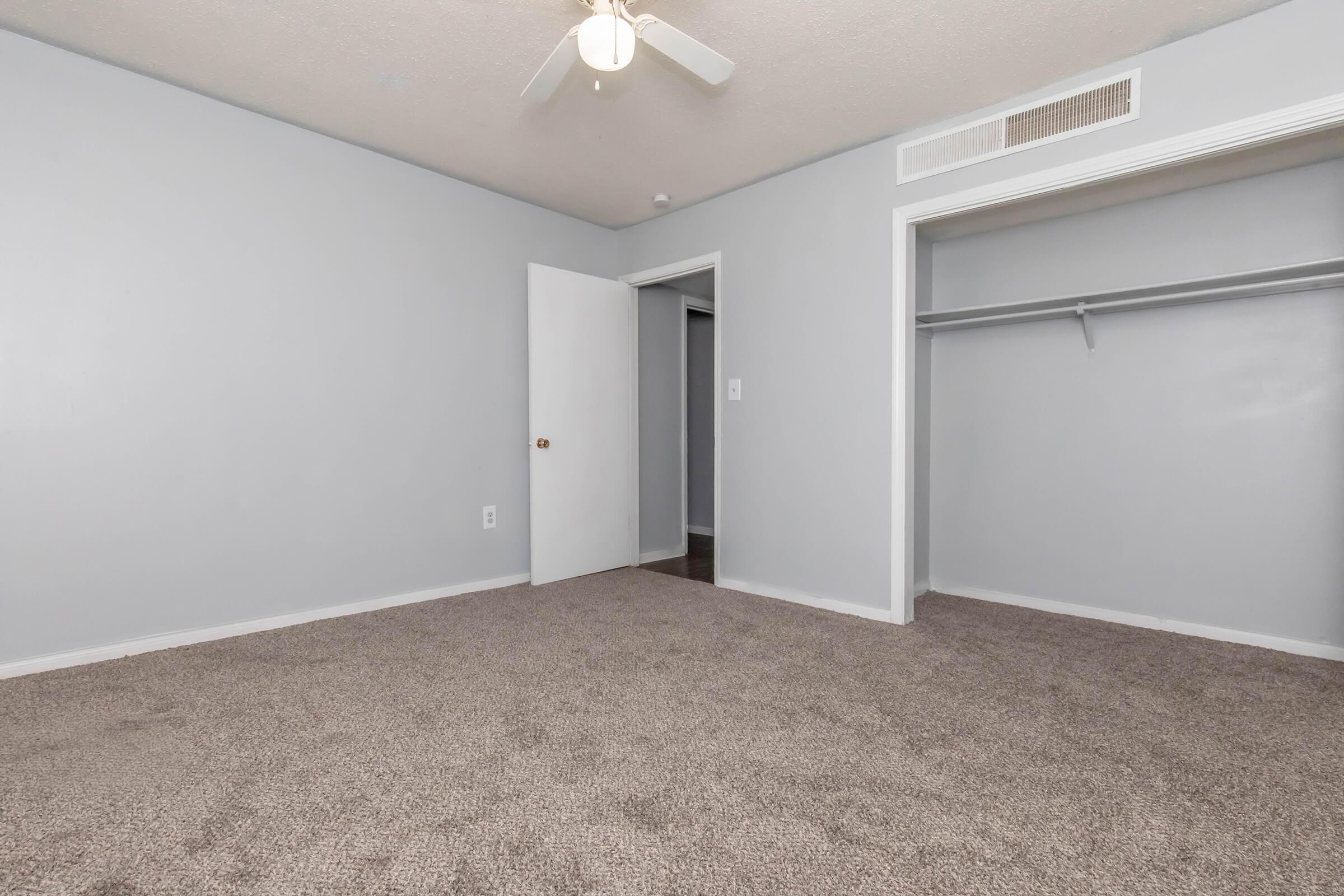 A small, empty room featuring light gray walls and beige carpet. There is a ceiling fan and a door leading to another area. A closet section is visible on the right side, and the overall lighting suggests a neutral, well-maintained space.