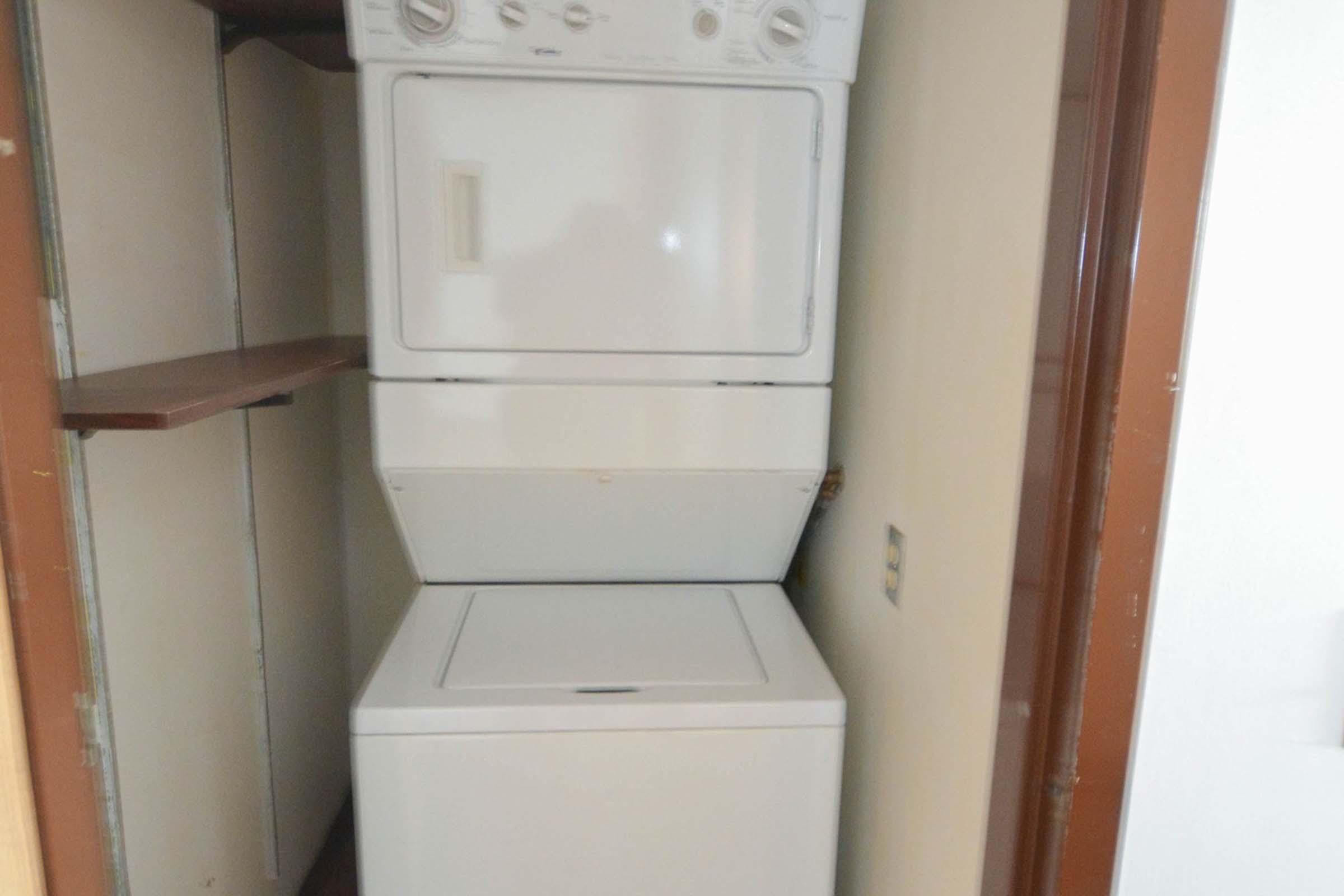 A stacked washer and dryer unit located in a closet space. The dryer is positioned on top of the washing machine, with some shelving above and on the side. The room appears to be well-lit and has a neutral-colored wall.