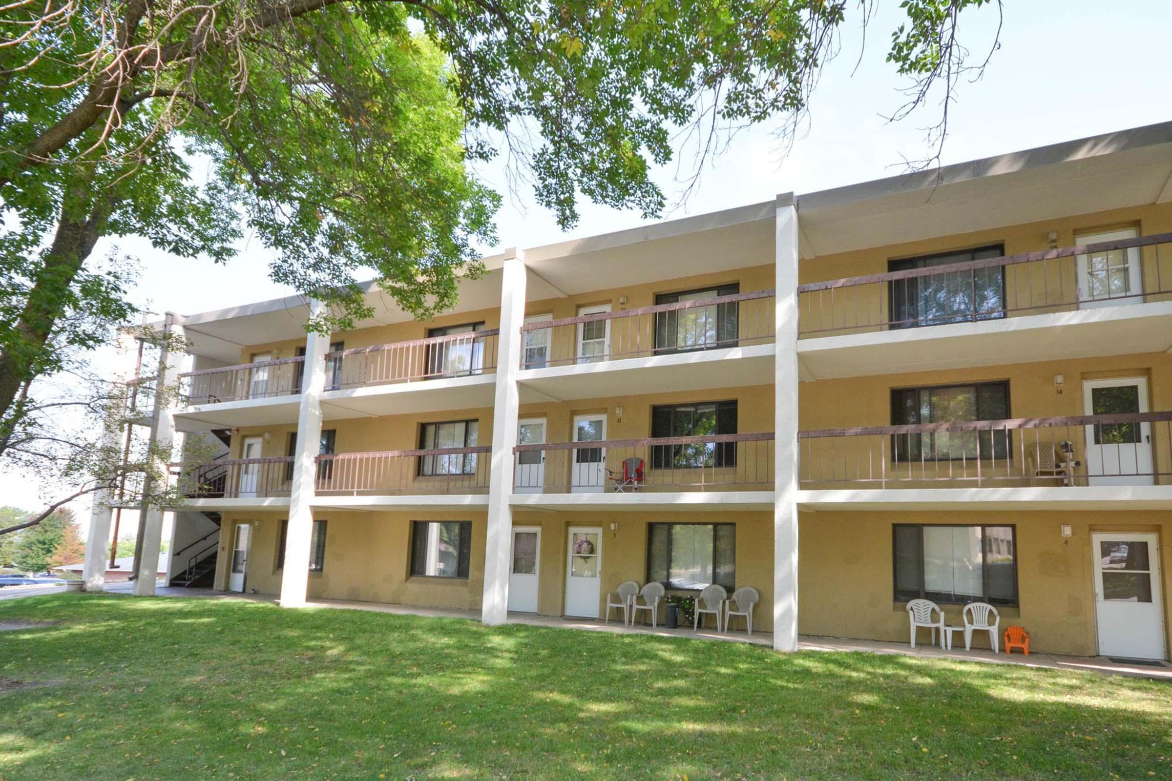 A multistory apartment building with a light-colored exterior, featuring several balconies and white doors. The building is surrounded by green grass and trees, with a few outdoor chairs visible on the lawn. The scene is bright and inviting, suggesting a residential neighborhood.