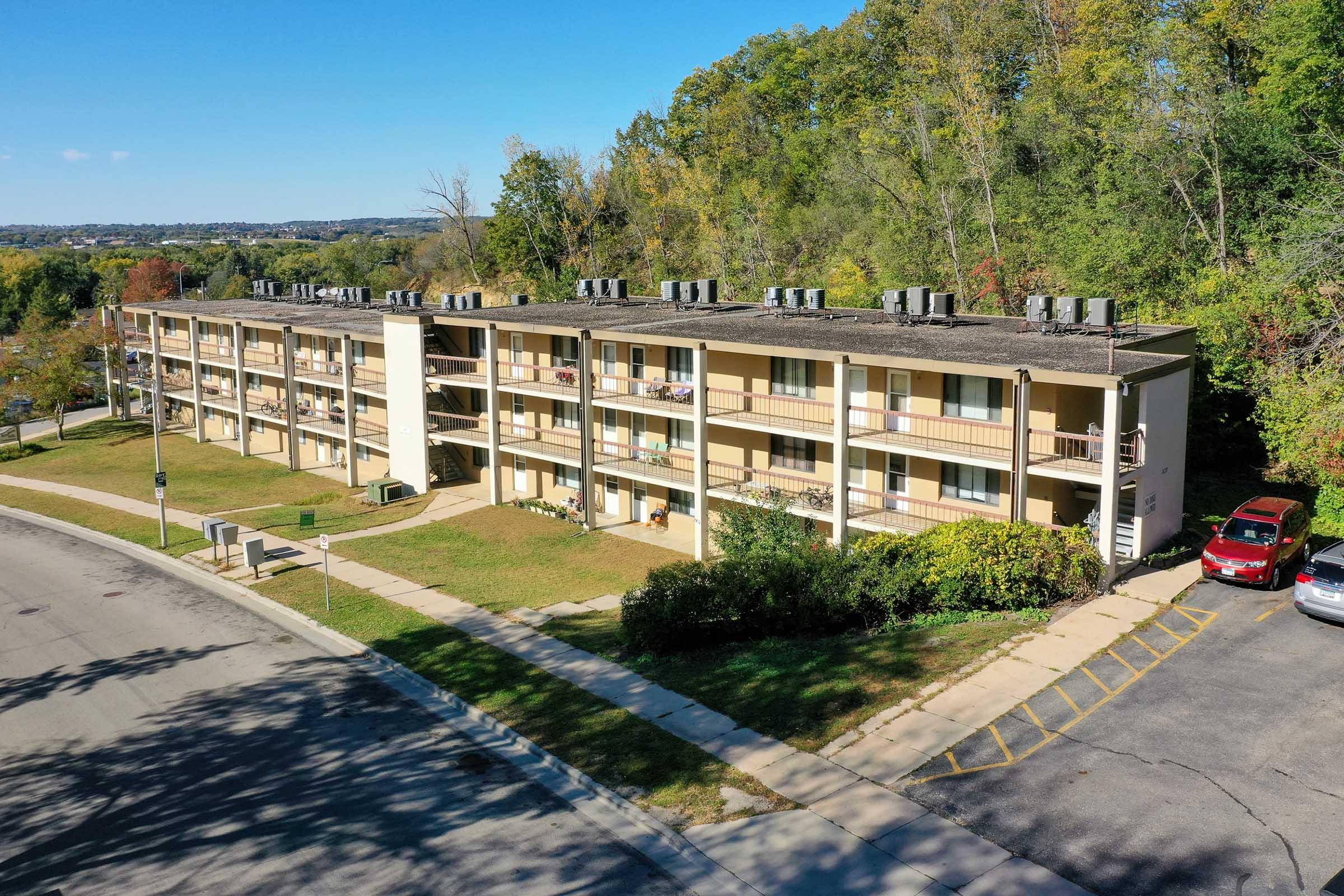 Aerial view of a three-story apartment building surrounded by greenery. The building features multiple balconies and air conditioning units on the roof. A parking lot with several cars is visible in front, along with a tree-lined background under a clear blue sky.