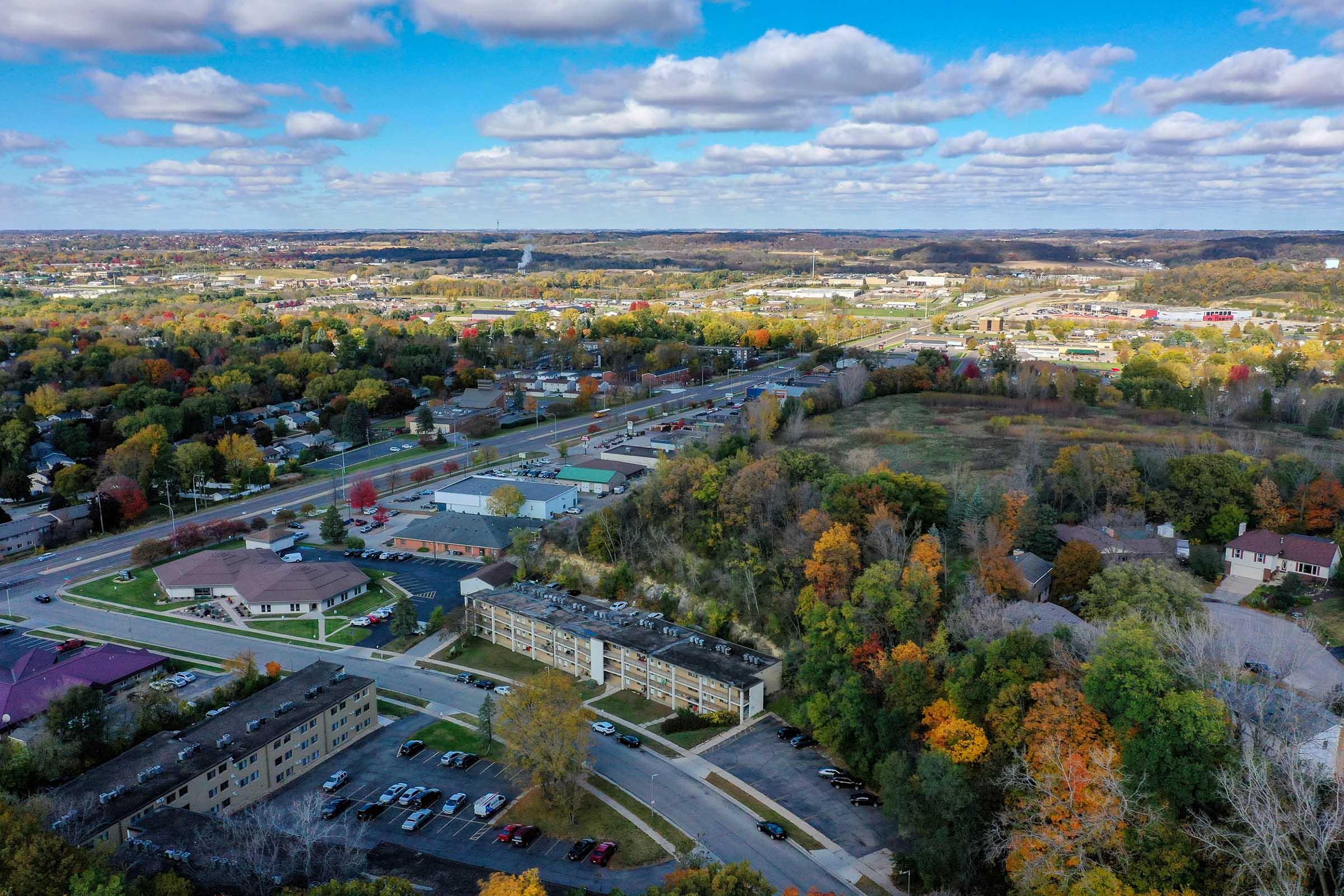 Aerial view of a suburban area showcasing a mix of residential buildings and commercial spaces. The landscape features autumn foliage with vibrant colors and a clear blue sky dotted with fluffy clouds. Roads and parking lots are visible, along with a patch of green land at the edge of the urban development.