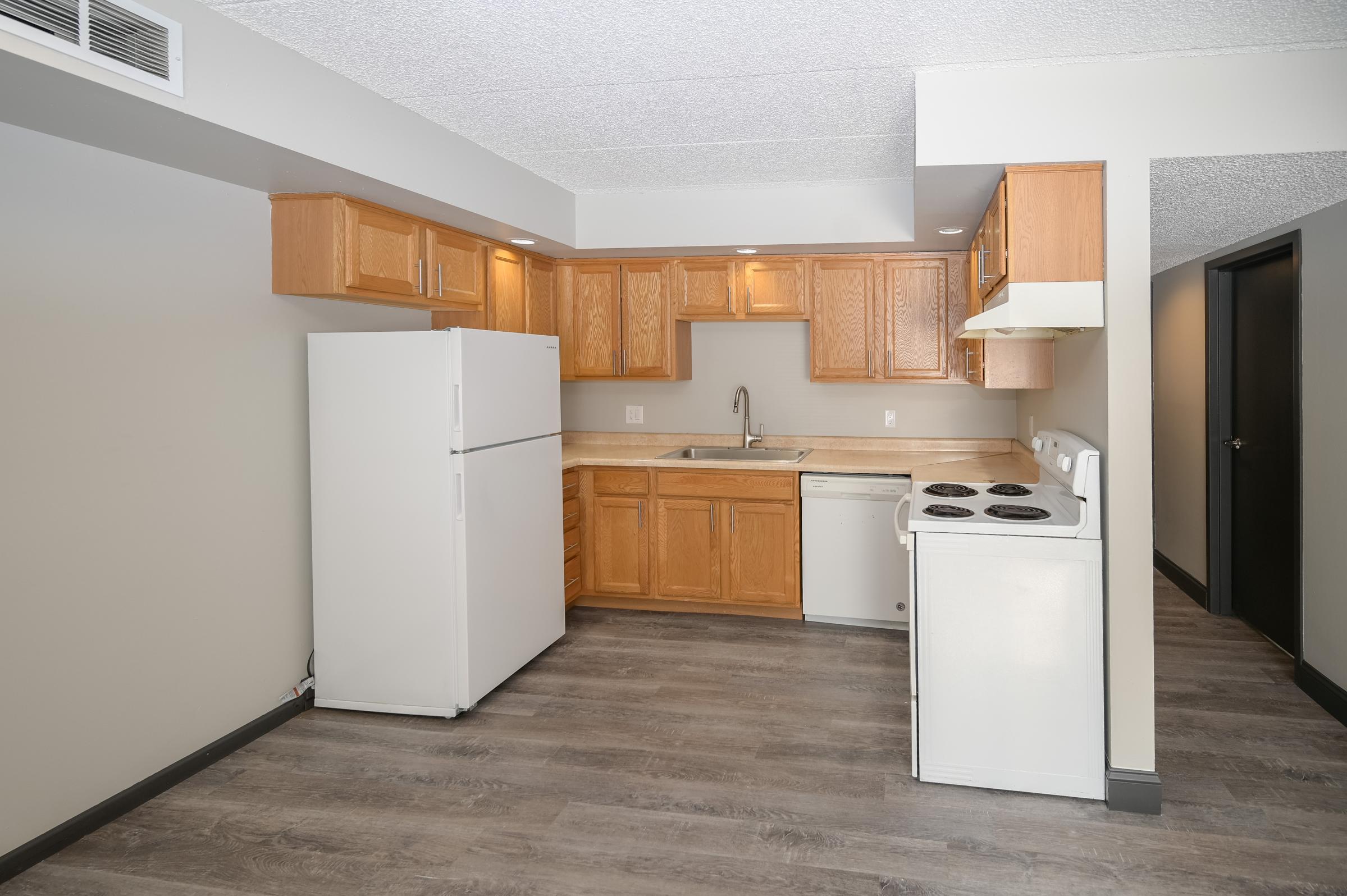 A modern kitchen featuring wooden cabinets, a white refrigerator, a sink, and a stove with an oven. The floor is covered with gray laminate, and the walls are painted in neutral tones. Adequate lighting is provided by overhead fixtures, creating a bright and spacious environment.