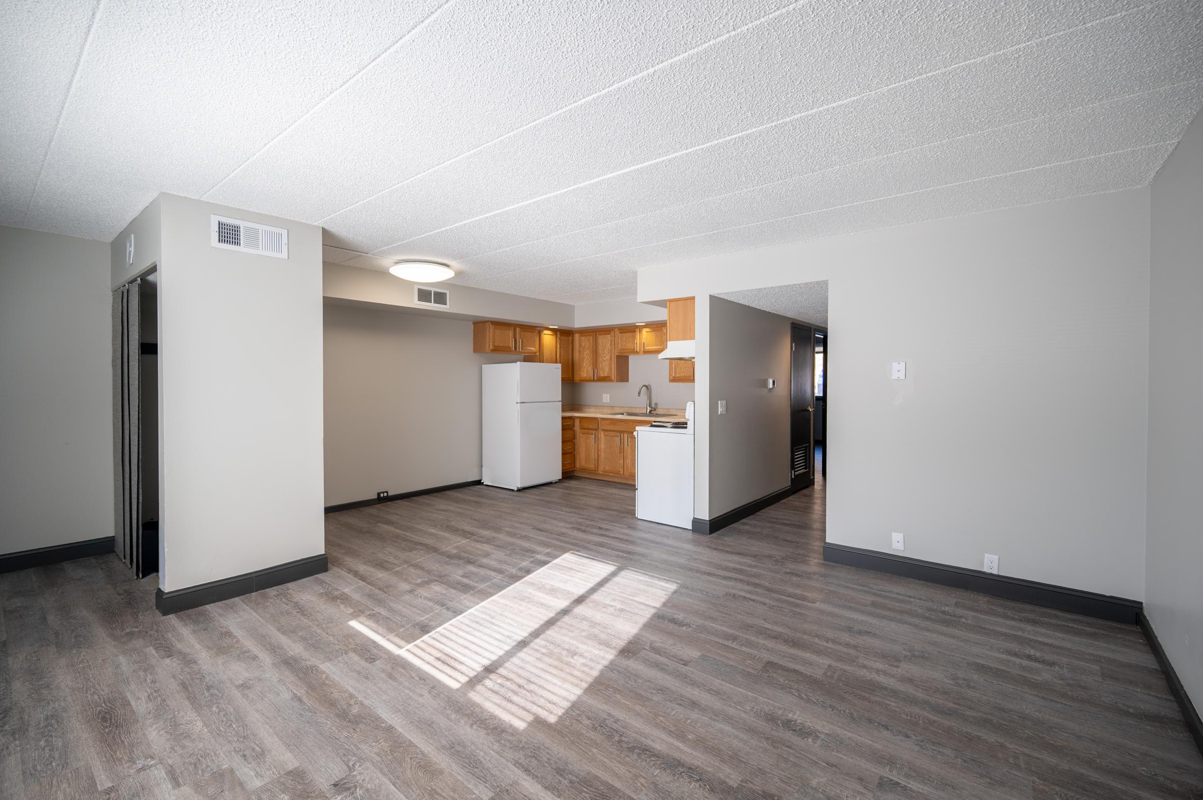 A spacious, unfurnished apartment interior featuring a light-colored wall, wooden cabinets, and a small kitchen area. The floor is covered with laminate flooring, and there is ample natural light coming in from a window. The layout includes an open space suitable for living and dining.