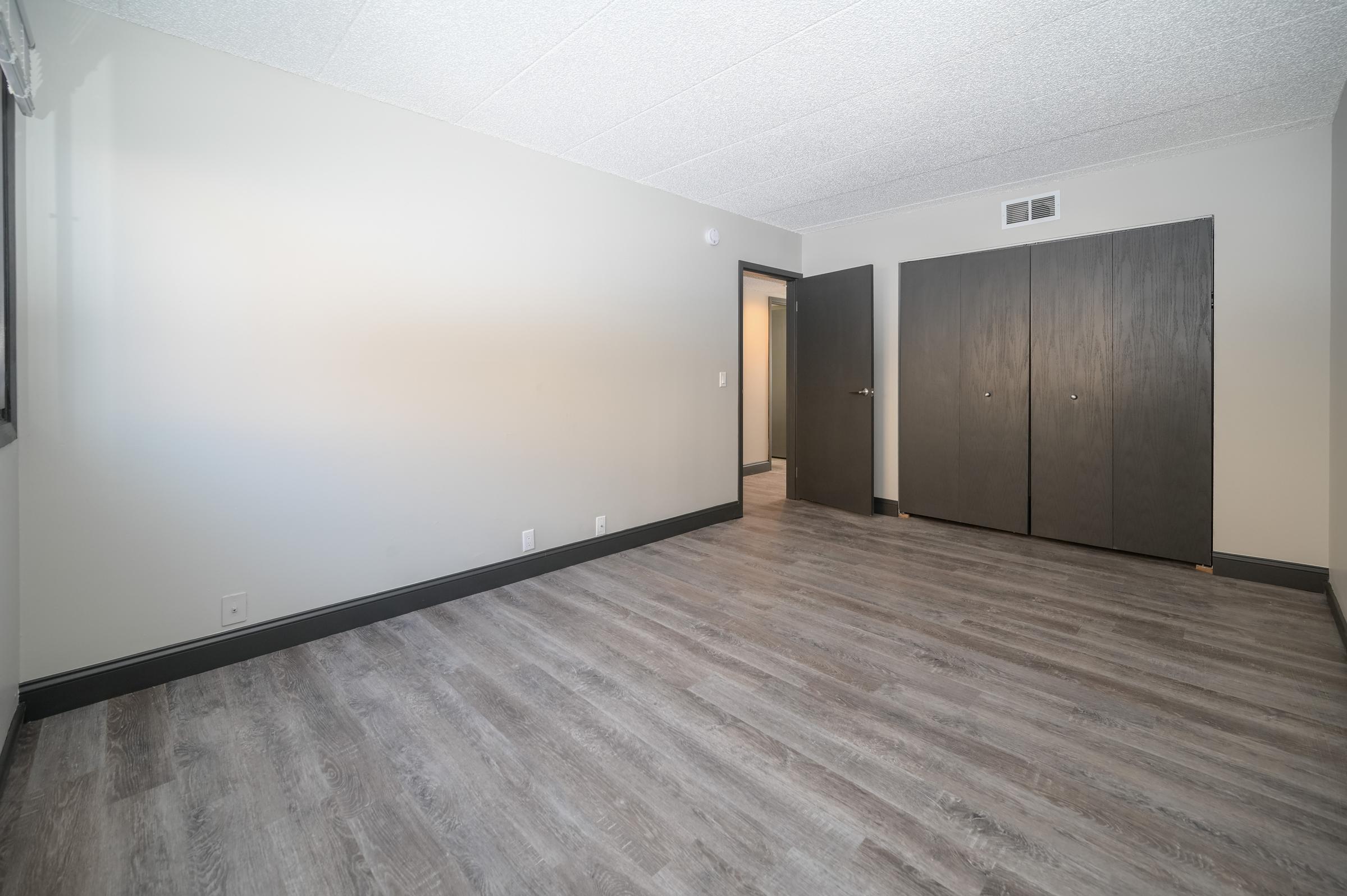 A spacious, vacant room with light gray walls and laminate flooring. Two large dark wood closet doors are on one side, and a doorway leads to another room. Natural light enters through a window, creating a bright and open atmosphere.