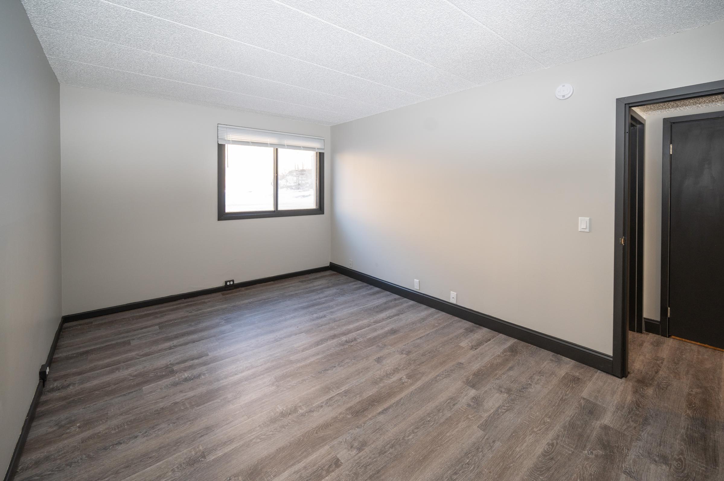 A vacant room with light gray walls and dark trim. The floor is covered in wood-like laminate. A single window allows natural light, and there are no furnishings. A door is visible on the right side, suggesting access to another space. The overall atmosphere is minimalist and open.