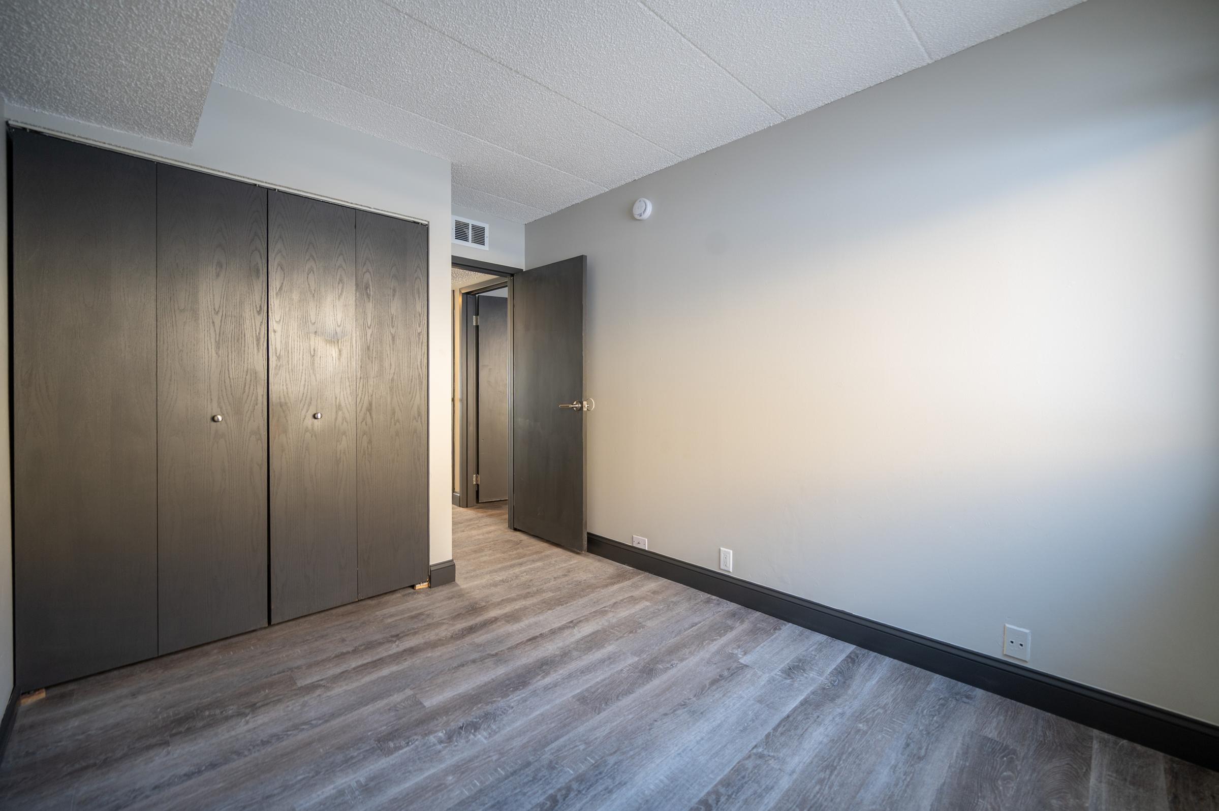 Interior view of a room featuring light-colored walls and dark wooden closet doors. A door leads to another space, and the flooring is a light wood finish. The room is sparsely decorated, creating a clean and open atmosphere.