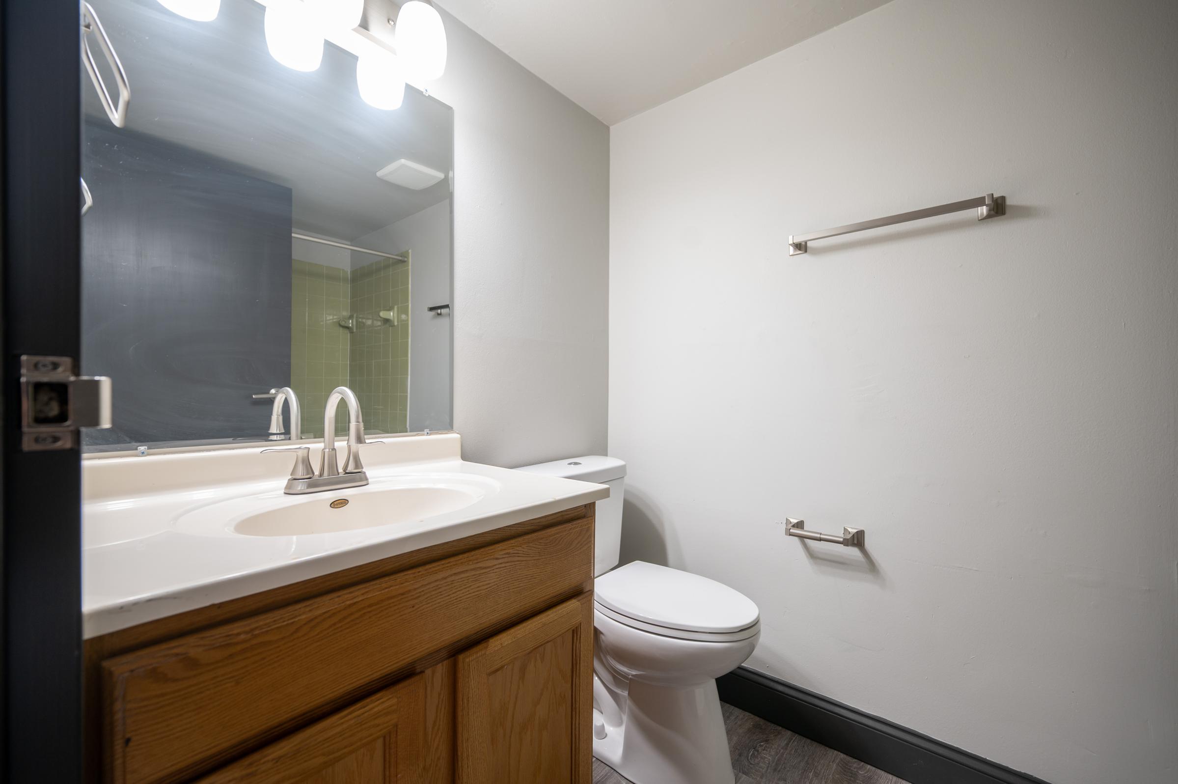 A clean bathroom featuring a white countertop with a sink, a mirror above, a toilet, and a towel rack. The walls are light gray, and the floor is dark. There is a shower area visible in the background with green tiles. Soft lighting is provided by overhead fixtures.