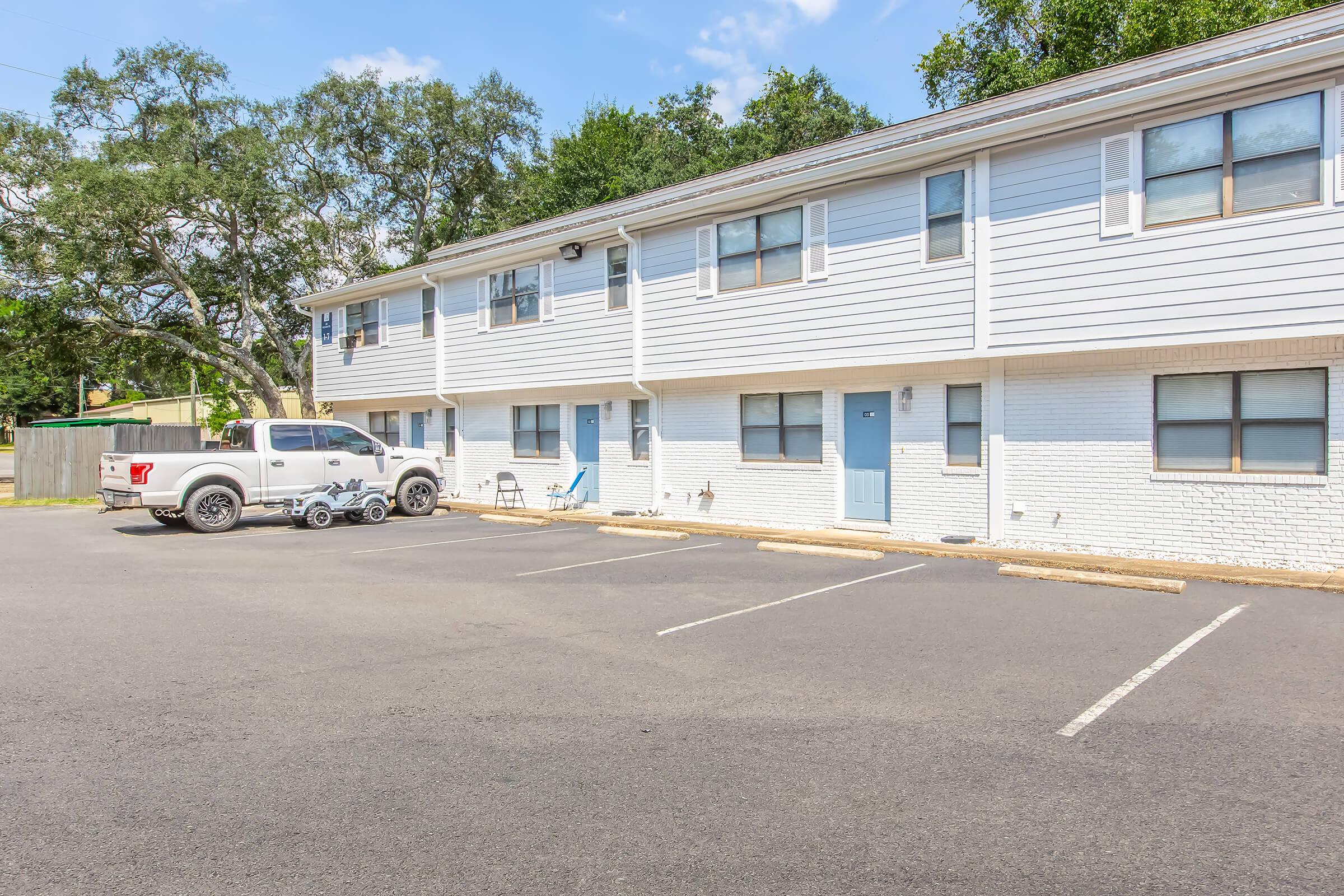 A row of two-story residential buildings with light blue siding and white brick accents. A parked white pickup truck is in the foreground, with a few empty parking spaces. The scene is set on a sunny day with clear blue skies and some greenery in the background.