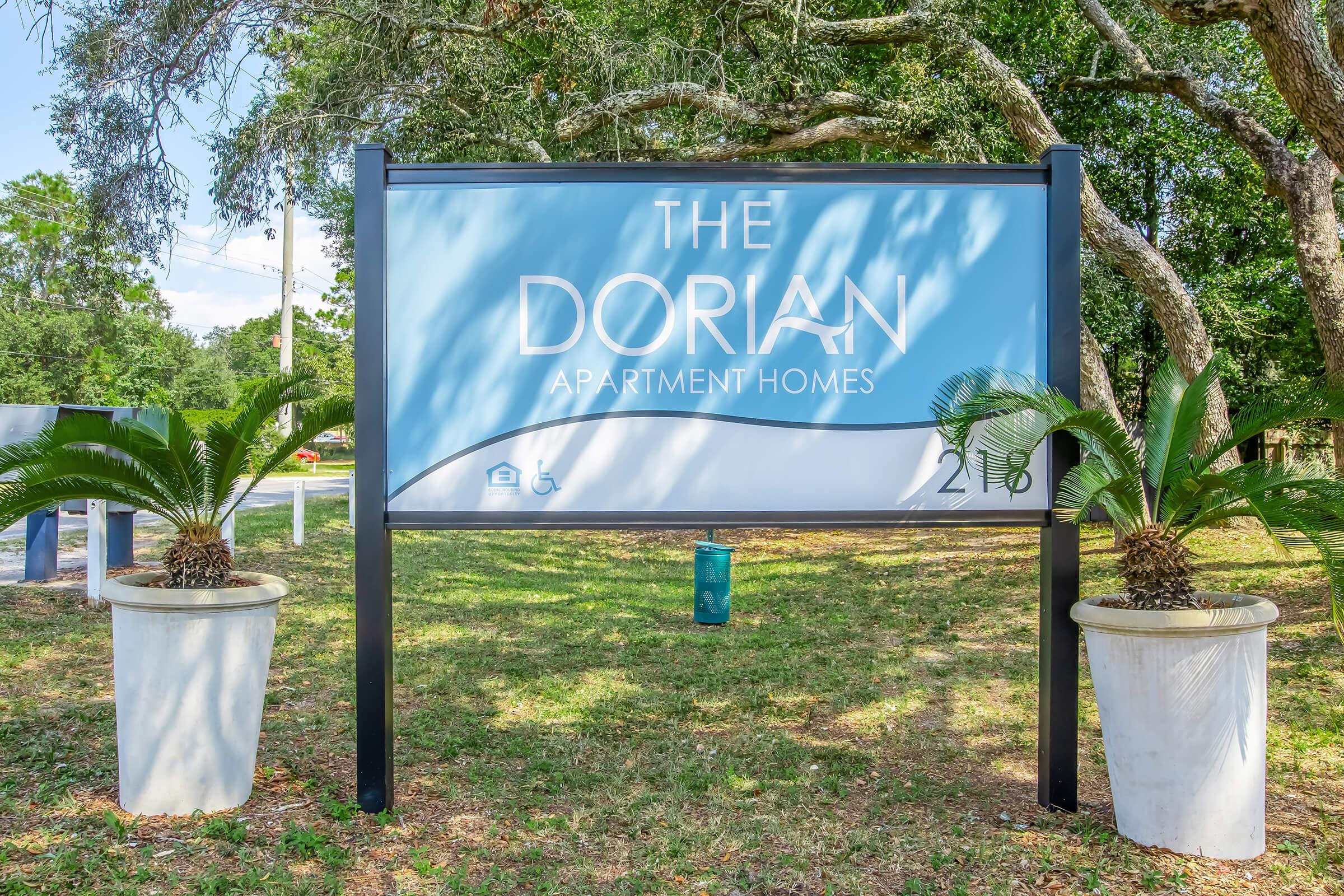 Sign for "The Dorian Apartment Homes" featuring a light blue background with white text. It is flanked by two planters with palm plants, set in a green landscape with trees and grass in the background. The sign includes a small icon indicating accessibility.