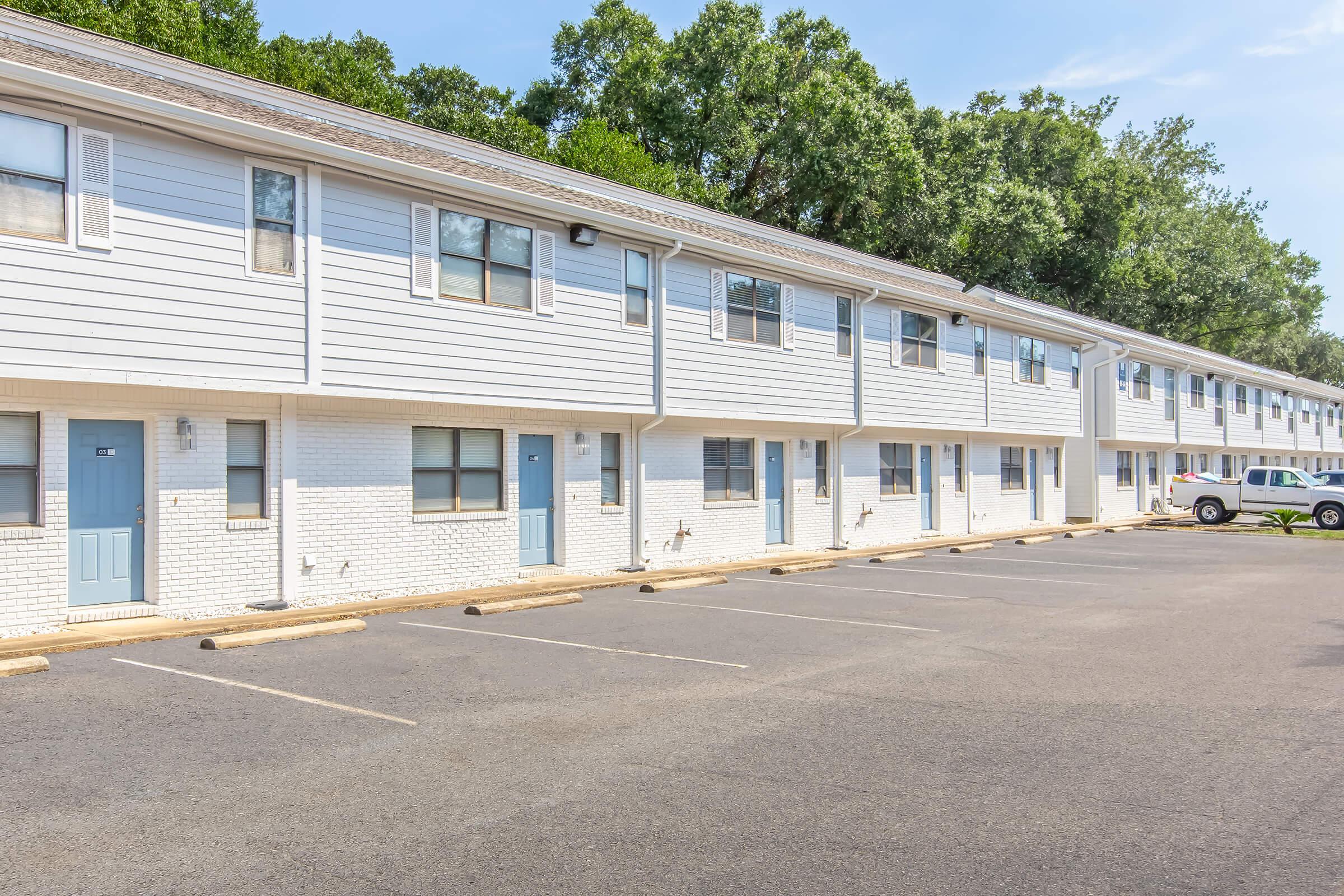 A row of two-story apartment buildings with light gray siding and white brick accents. Each unit has a blue door, and there are several parking spaces in front. Lush green trees line the background under a clear blue sky, creating a pleasant residential setting.