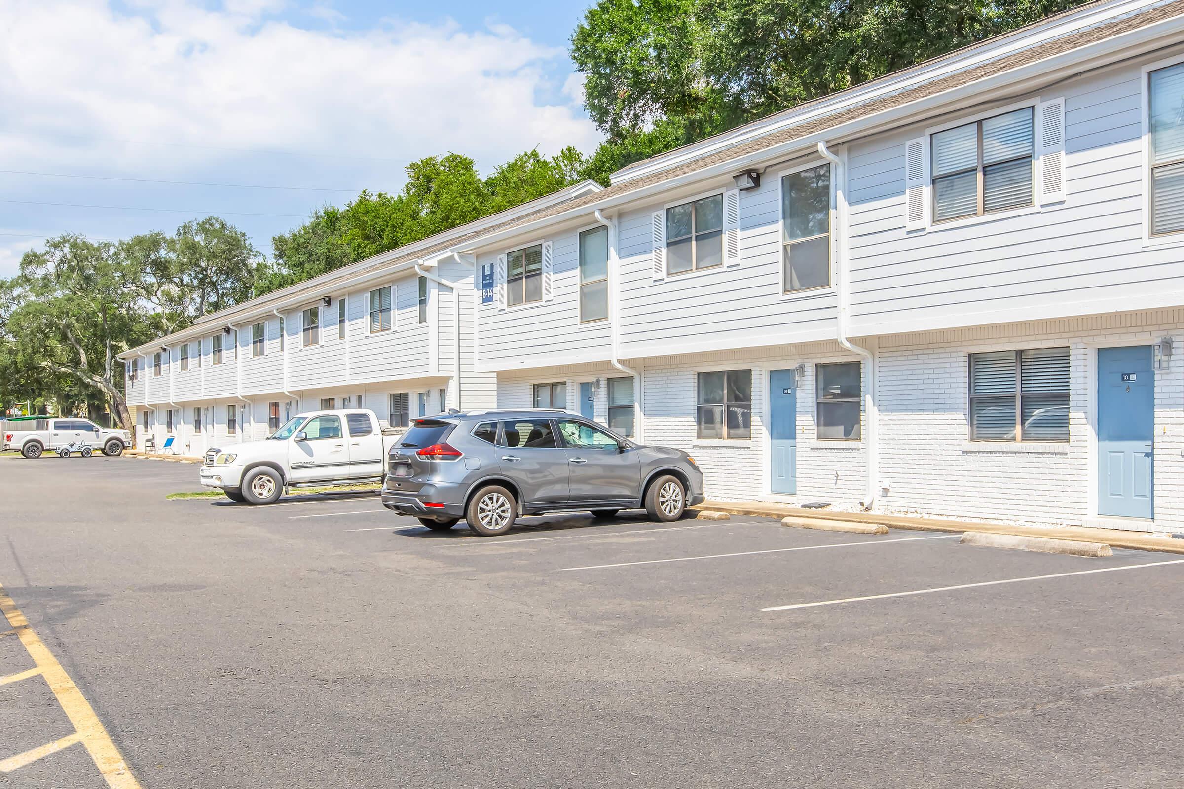 A row of two-story apartment buildings with light gray siding, featuring multiple units. Each unit has a blue door and several windows. In the foreground, parked cars, including a gray SUV and a white vehicle, are visible on a paved parking lot. Lush green trees are in the background under a partly cloudy sky.