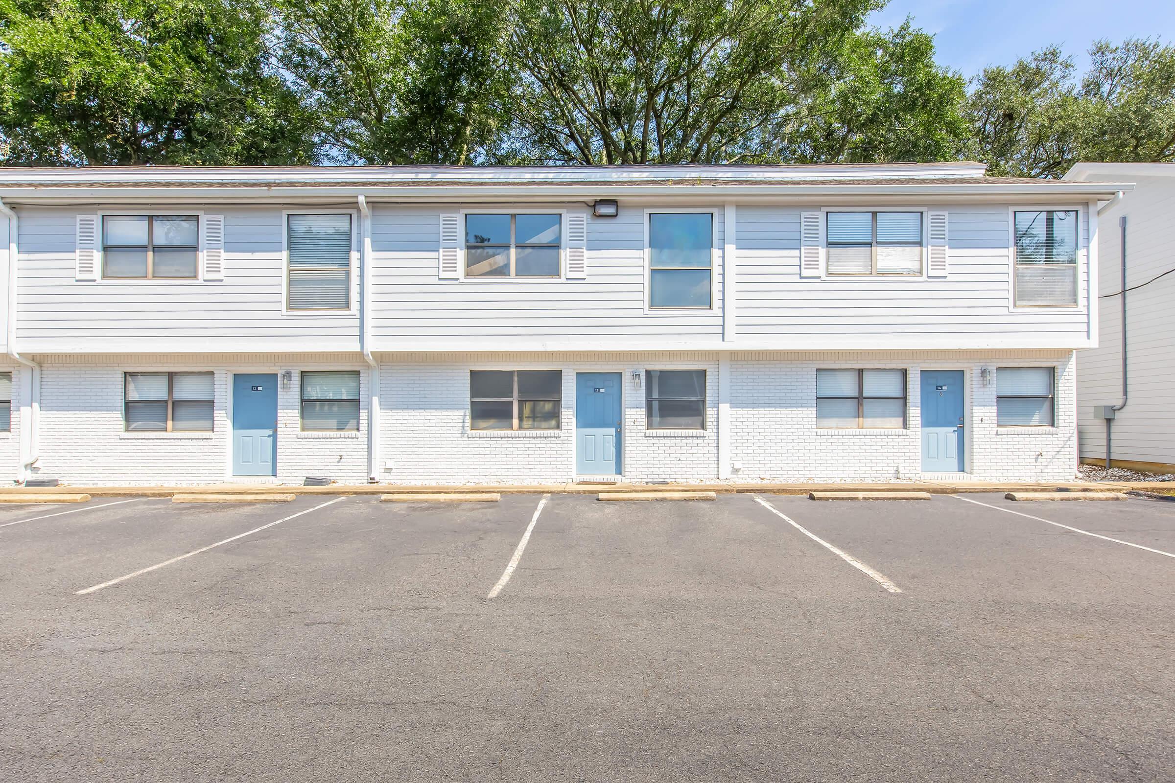 A two-story building with a modern design, featuring white siding and blue doors. There are several parking spaces in front of the building, with trees visible in the background under a clear blue sky. The structure is well-maintained and has multiple windows.