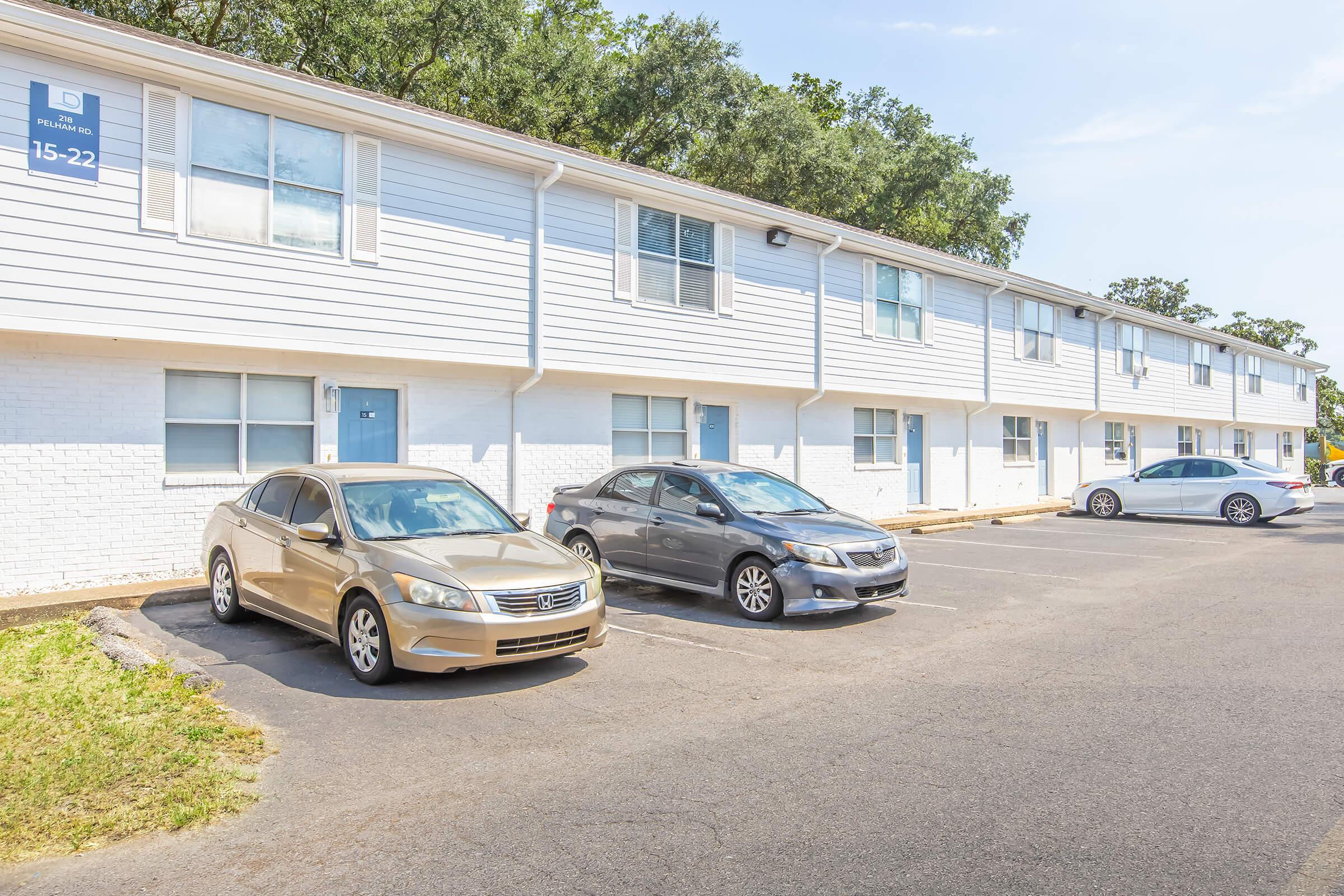 A row of two-story apartment units features several parked cars in front. The building has white siding and multiple windows, with a clear blue sky overhead. A few trees are visible in the background, adding greenery to the scene.
