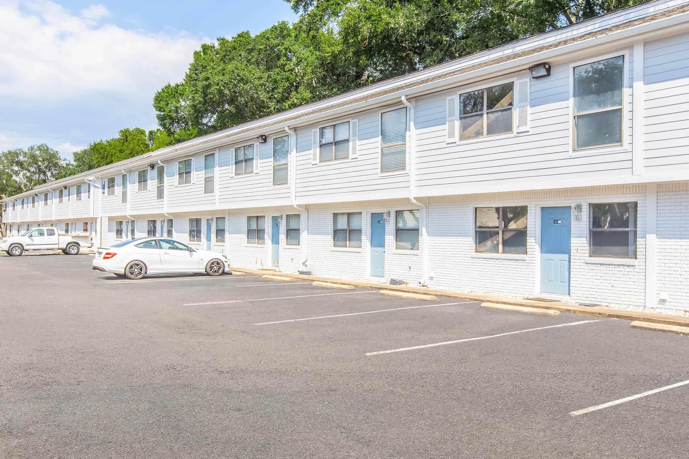 Row of white multifamily housing units with blue doors, set against a backdrop of greenery. Several parking spaces are visible in front, with a white car parked. The sky is partly cloudy, indicating a bright day.