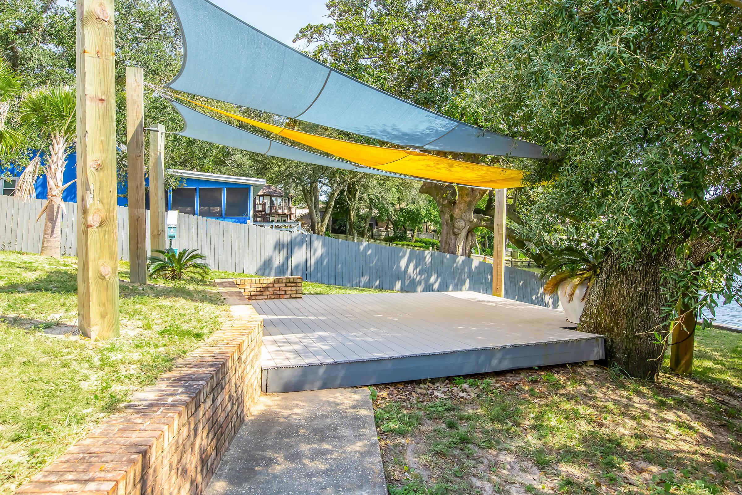 A shaded outdoor deck area near a lake, featuring a wooden platform with yellow and blue fabric awnings above. Surrounding greenery includes trees and shrubs, with a brick border along the pathway leading to the deck. A blue house is visible in the background, partially obscured by trees.