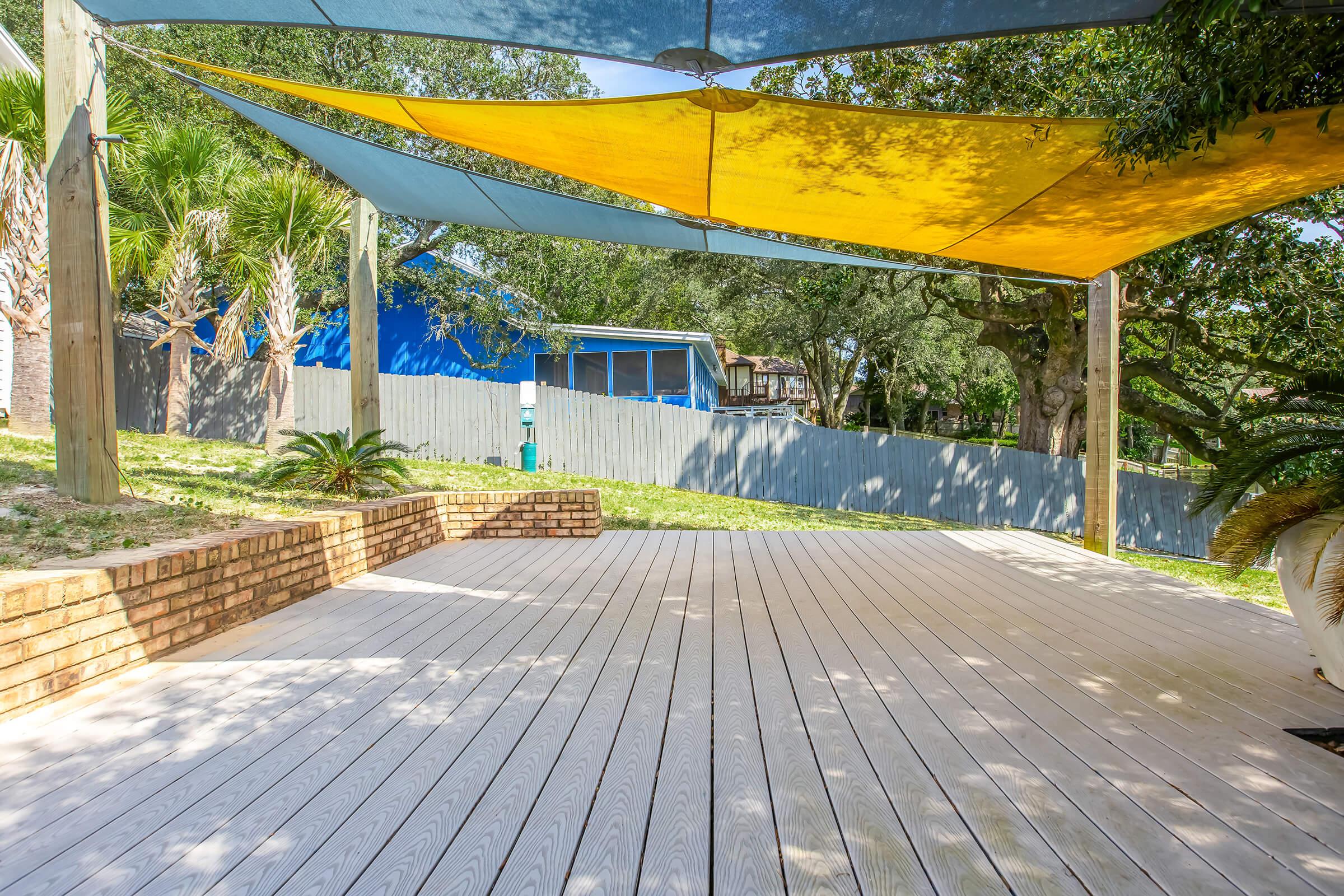 A view of a wooden deck with a colorful shade sail overhead, set in a green outdoor space. In the background, there are trees and a blue building behind a wooden fence, creating a relaxed, sunny atmosphere.