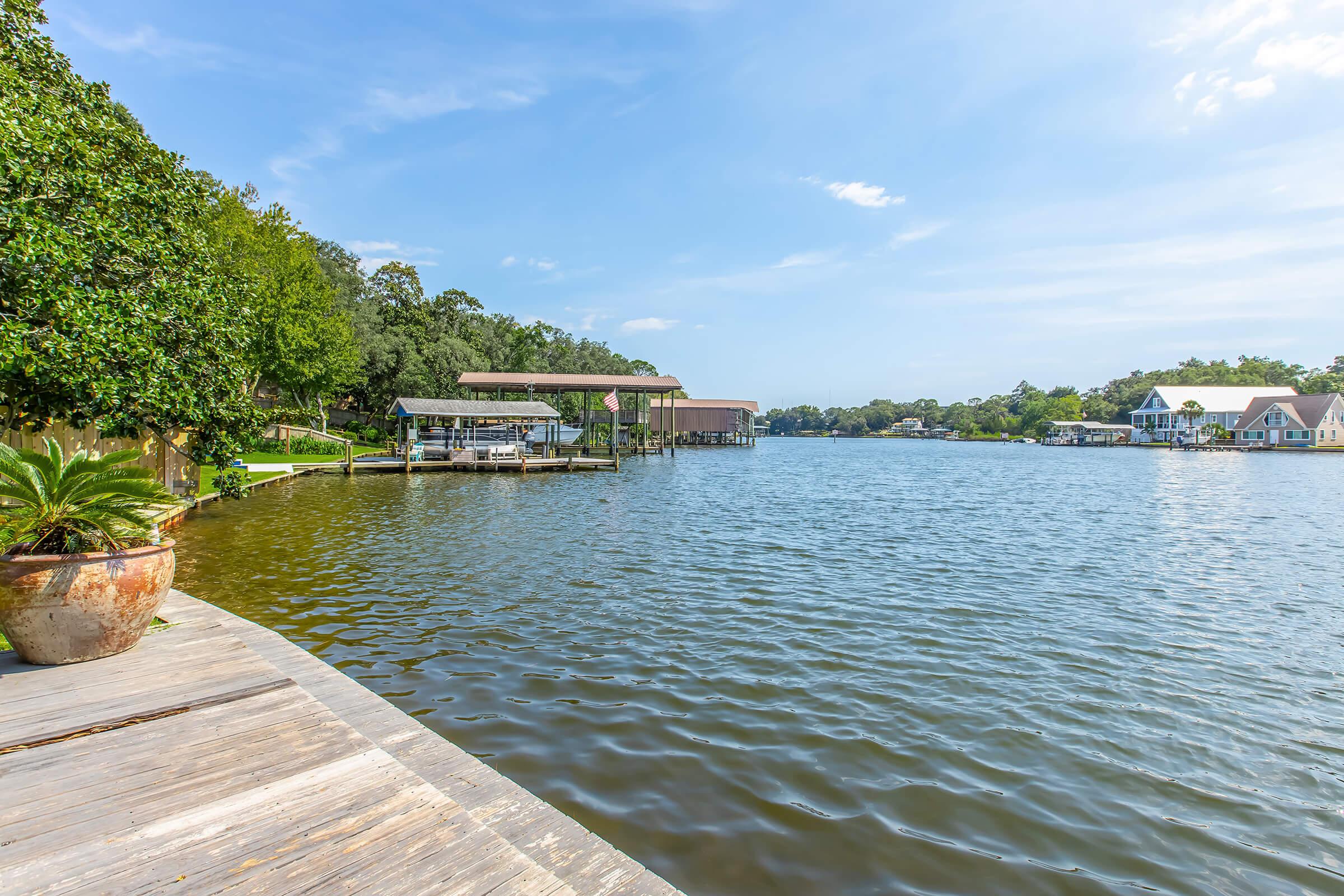 A peaceful riverside view featuring calm water, a wooden dock in the foreground, and lush greenery on the left. On the right, picturesque houses with boat docks are visible along the shoreline under a blue sky with scattered clouds.