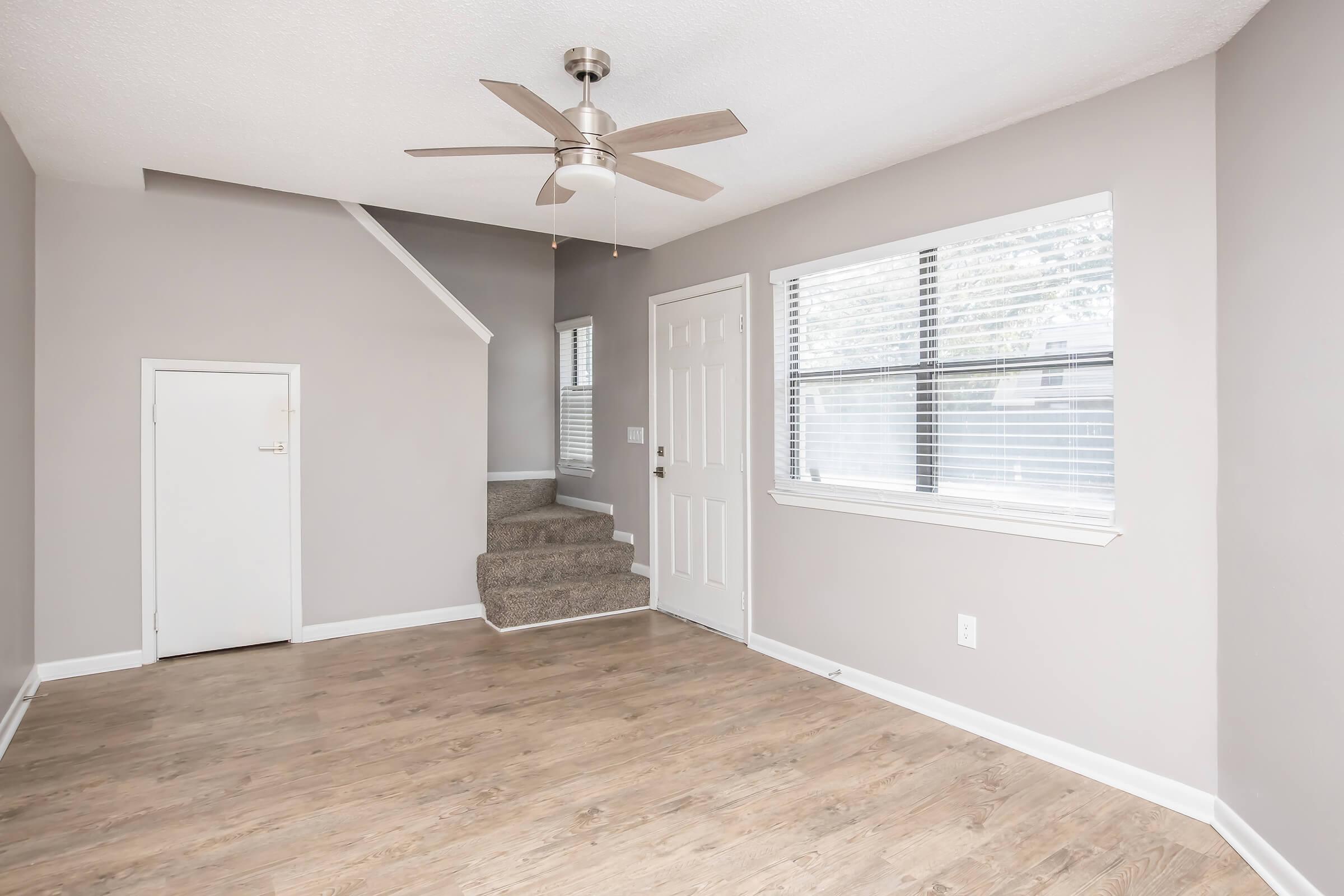A light and airy living space featuring beige walls and wood-like flooring. A ceiling fan is installed, and there are two windows allowing natural light. In the corner, there is a closed door, and to the side, a staircase leads to another level. The overall atmosphere is modern and inviting.