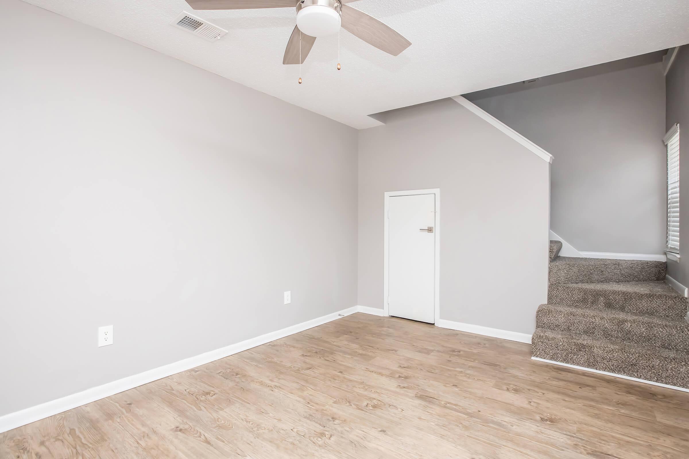A spacious, empty living room with light gray walls, a ceiling fan, and wooden flooring. A closed door is visible on one wall, and a staircase with carpet leads to an upper level. Natural light comes through a nearby window, creating a bright and inviting atmosphere.