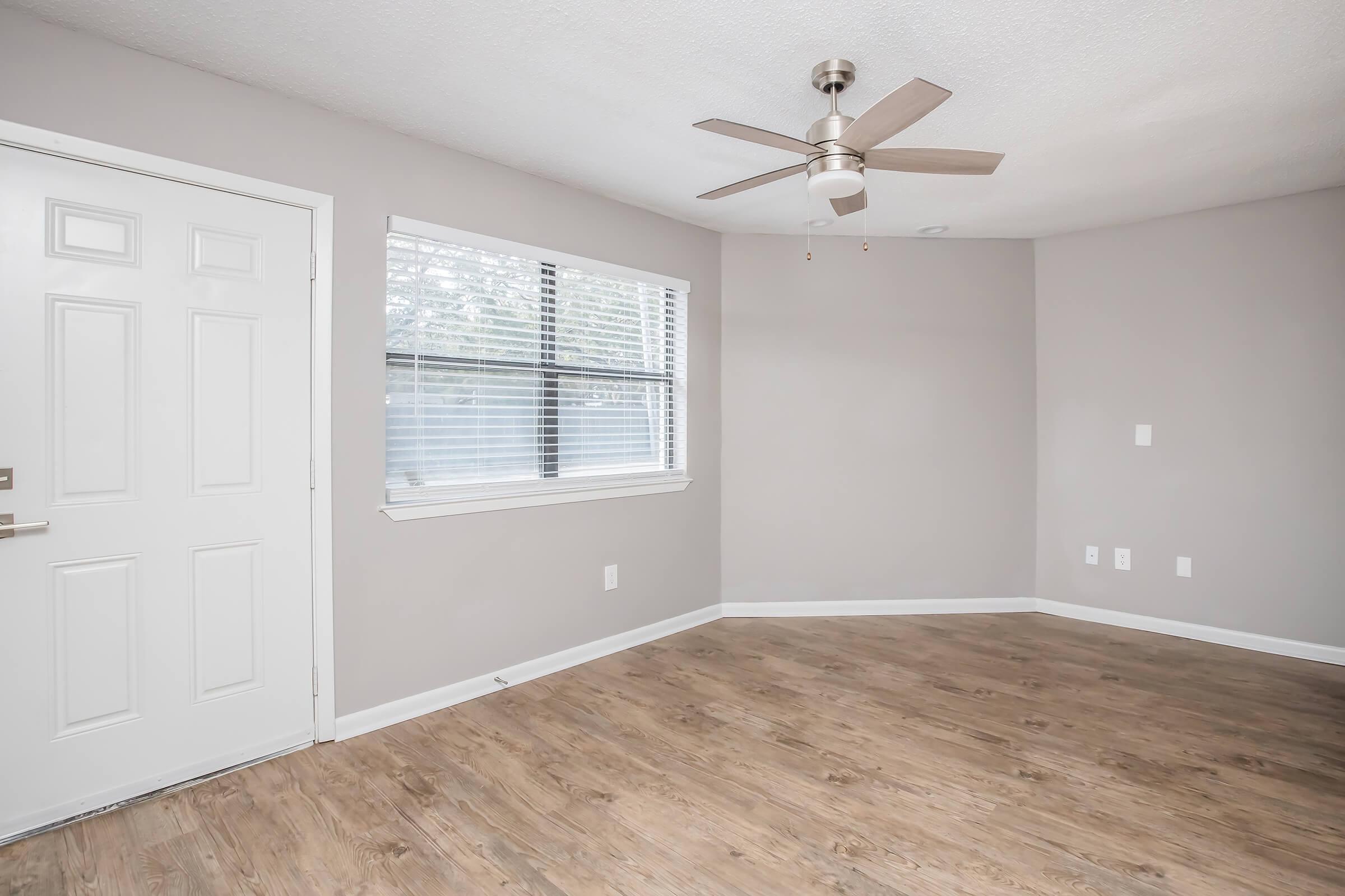 A vacant room featuring a ceiling fan, light-colored walls, and wood-style flooring. There is a white door on the left side, a window with blinds on the right, and minimalist decor, giving the space a clean and airy look.