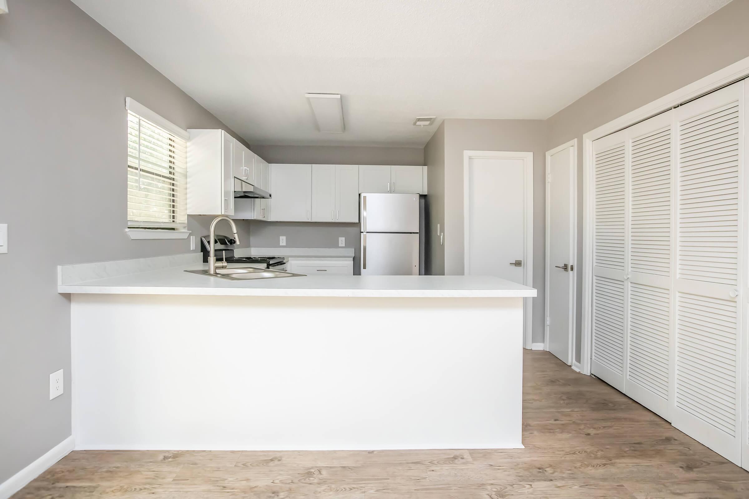 A modern kitchen featuring a white countertop island, stainless steel appliances, and bright cabinetry against a gray wall. Natural light streams through a window, illuminating the space. A door leads to a closet or pantry, enhancing the kitchen's functionality and contemporary design.